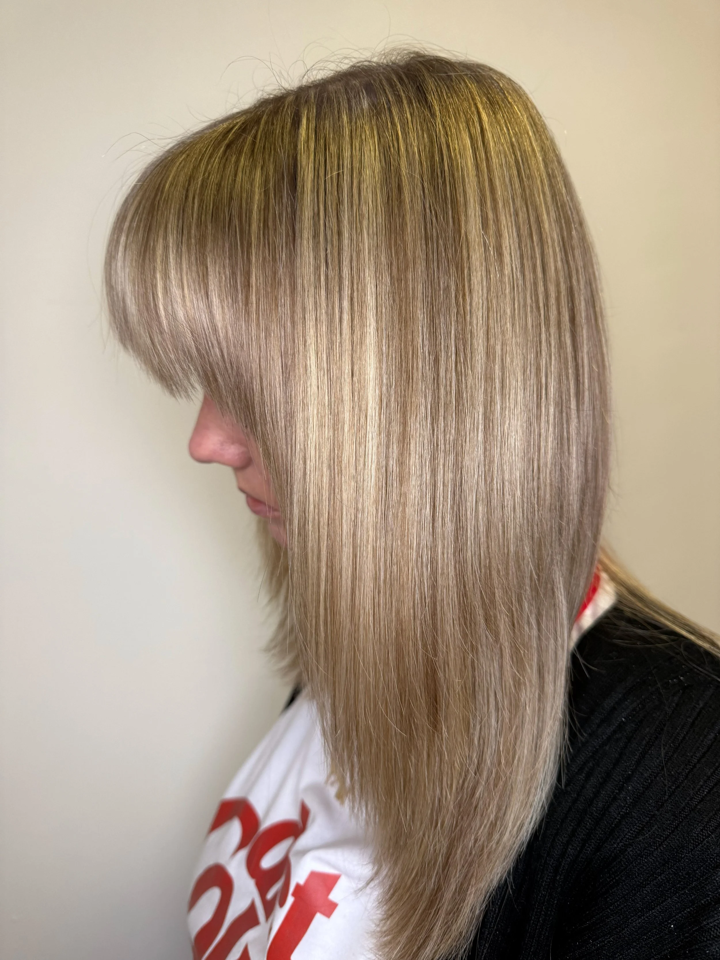 Side view of a woman with straight, blonde hair with darker roots, wearing a white shirt with red lettering and a black jacket.
