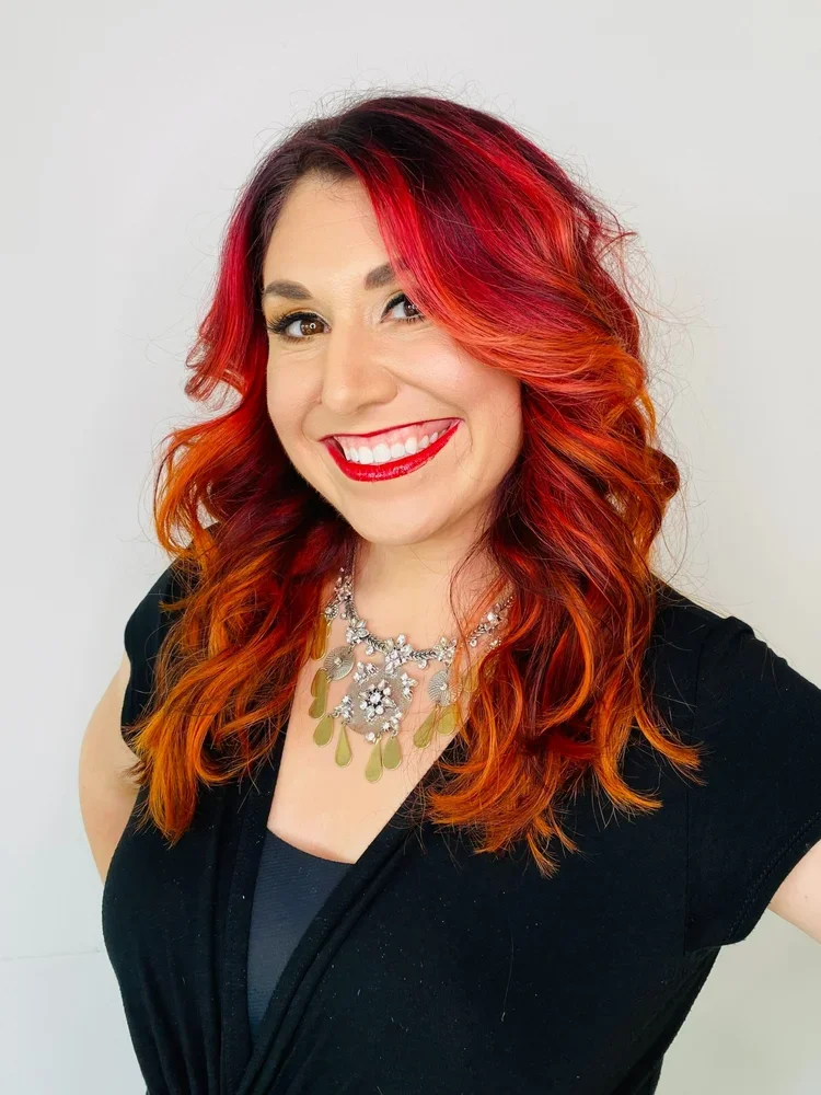 A woman with red and orange curly hair smiling, wearing red lipstick, a black top, and a statement necklace with silver and green embellishments.
