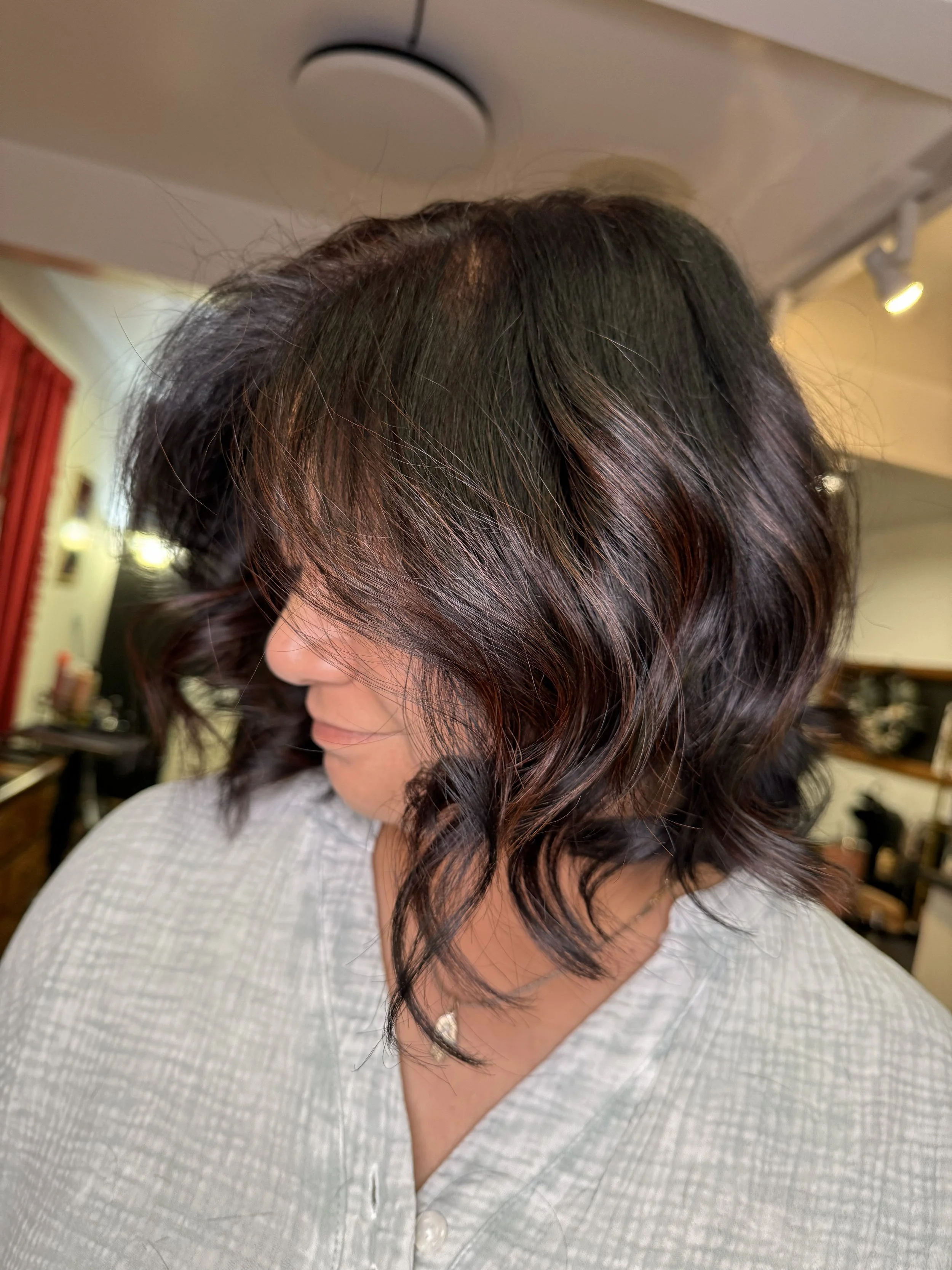 A woman with shoulder-length wavy dark hair with reddish highlights, wearing a light gray top and a necklace, with her face turned to the side in a salon or home setting.