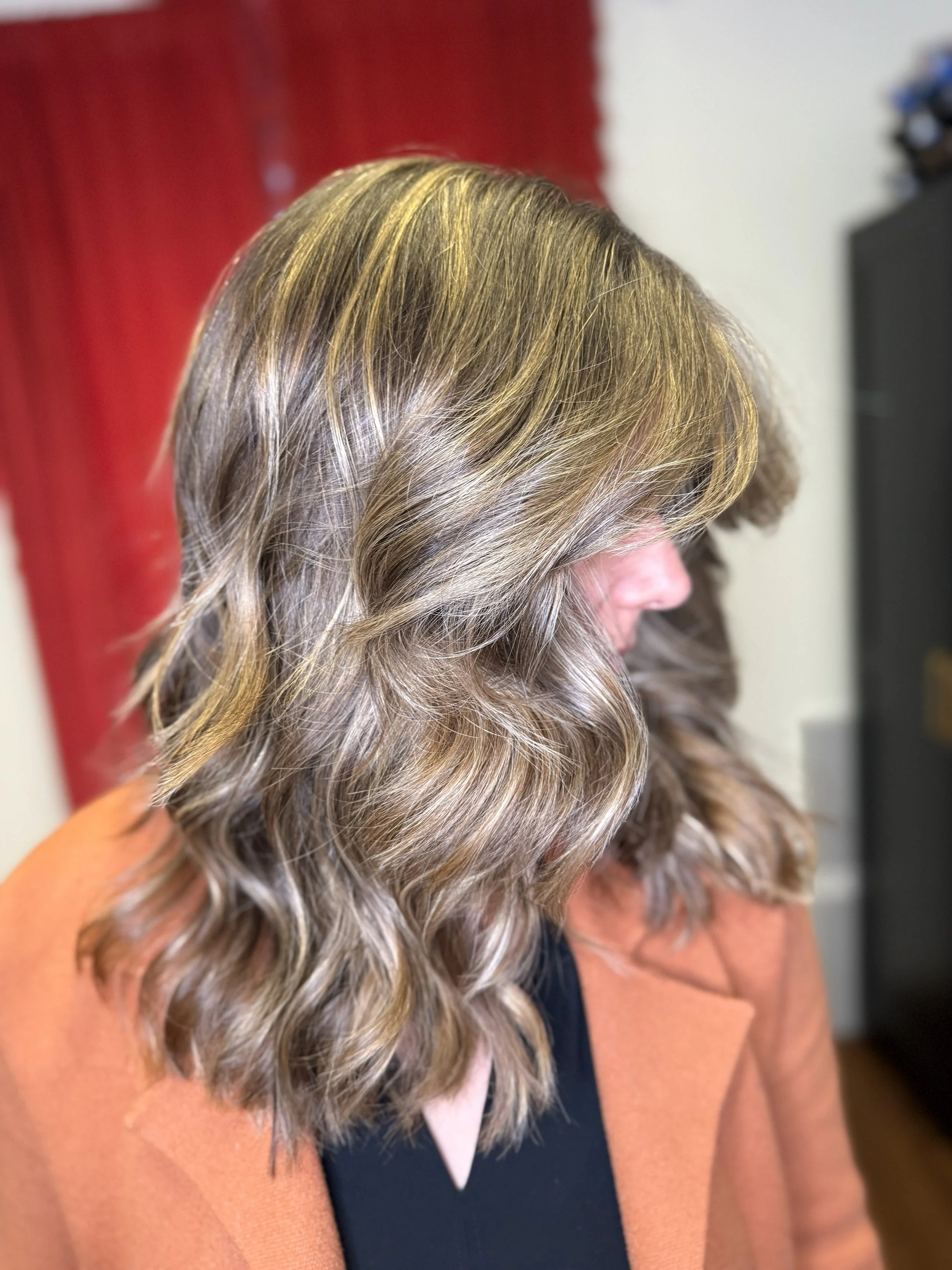 A woman with shoulder-length, wavy blonde hair, seen from the side, wearing a black top and a brown blazer.