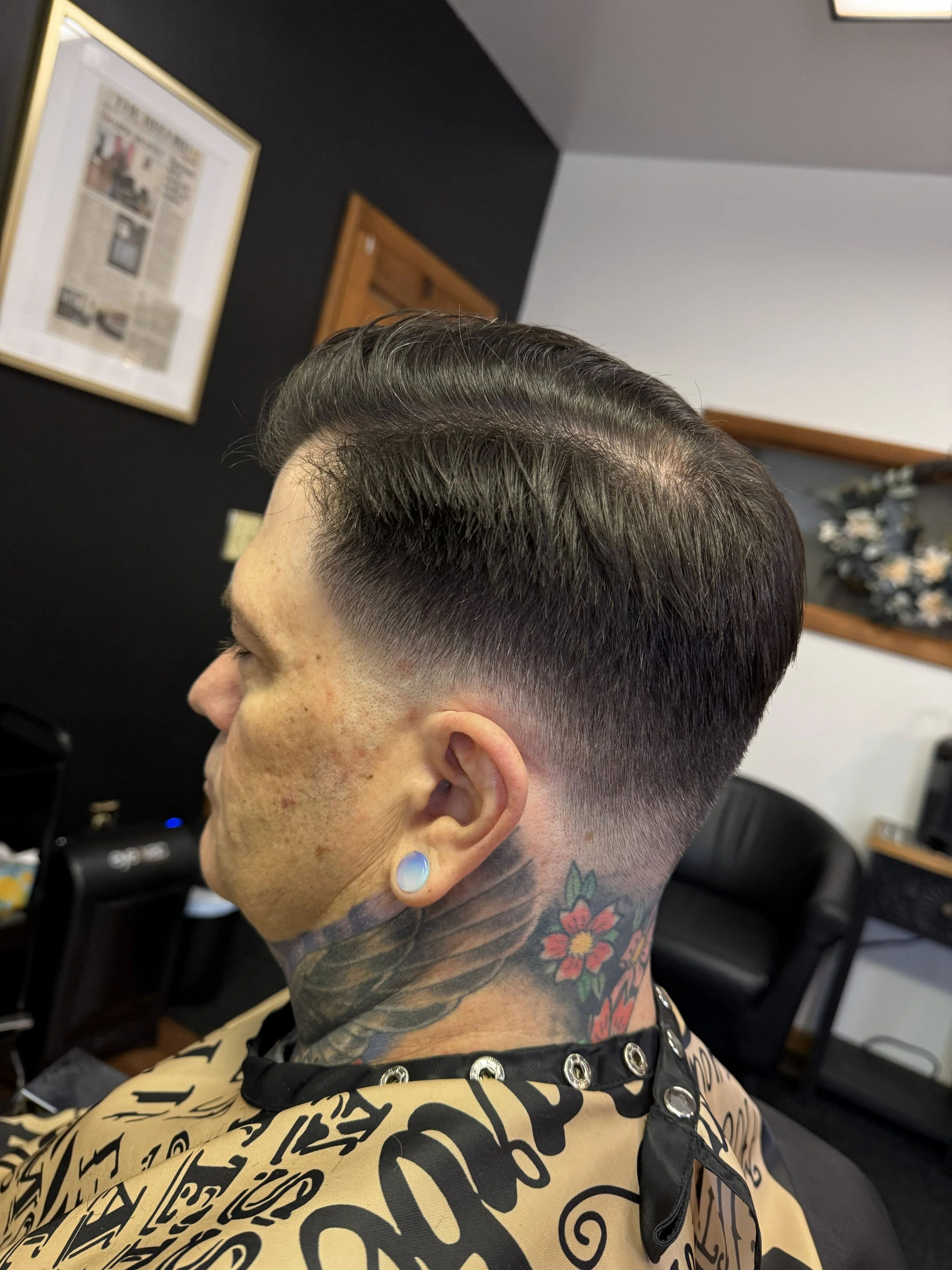 Man with a modern haircut featuring a skin fade on the sides and long, slicked-back hair on top, seated in a barbershop with a tattoo of a flower on his neck and a large gauge earlobe piercing.