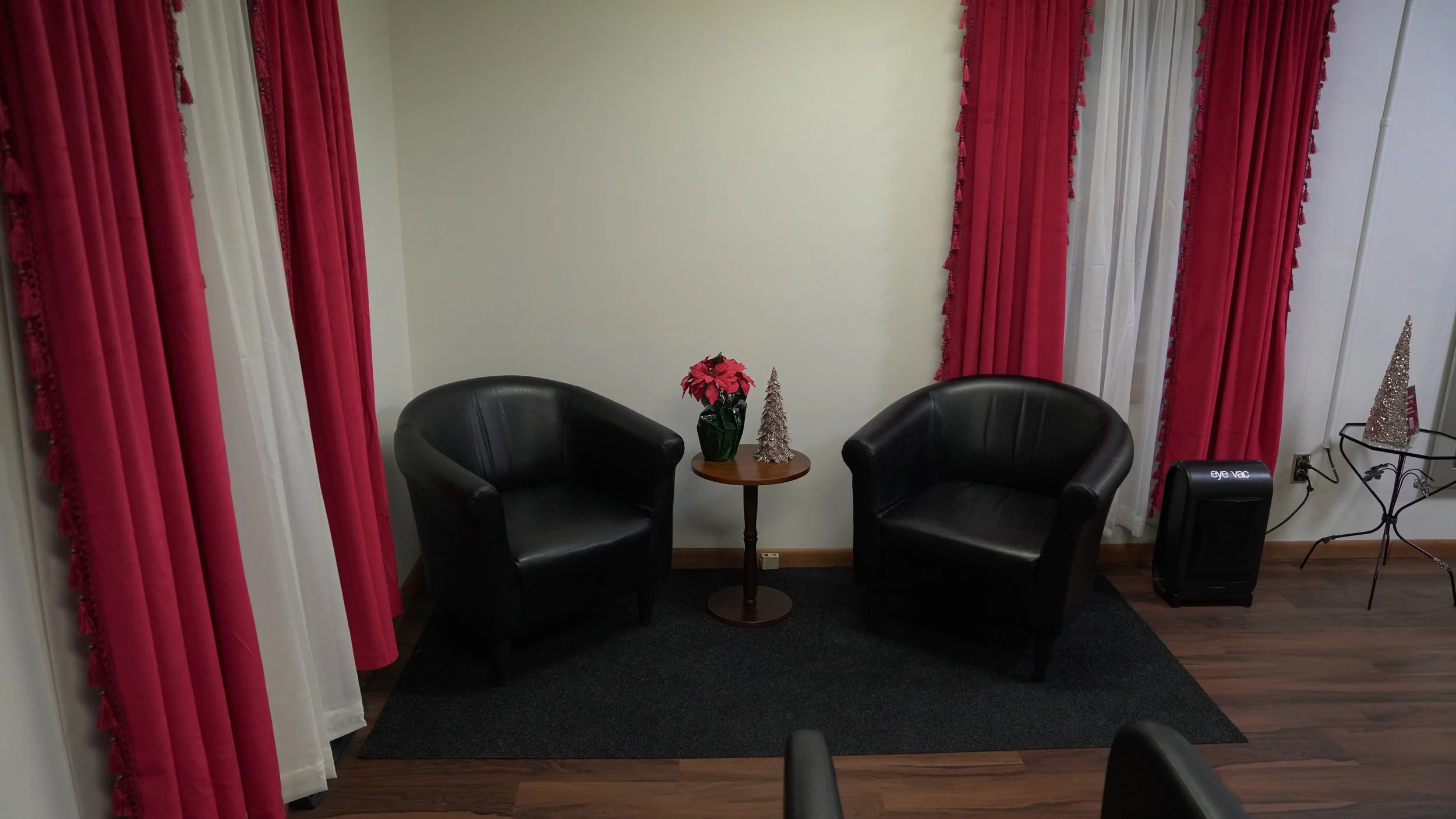 A cozy indoor setup with two black leather armchairs facing each other, separated by a small round wooden table. The table holds a vase with pink poinsettia flowers and two decorative silver Christmas trees. Red and white curtains with tassels adorn the window behind the chairs. The floor is wood with a small black rug underneath the chairs and table. To the right, there is a small black space heater and a decorative metal stand with a silver Christmas tree.