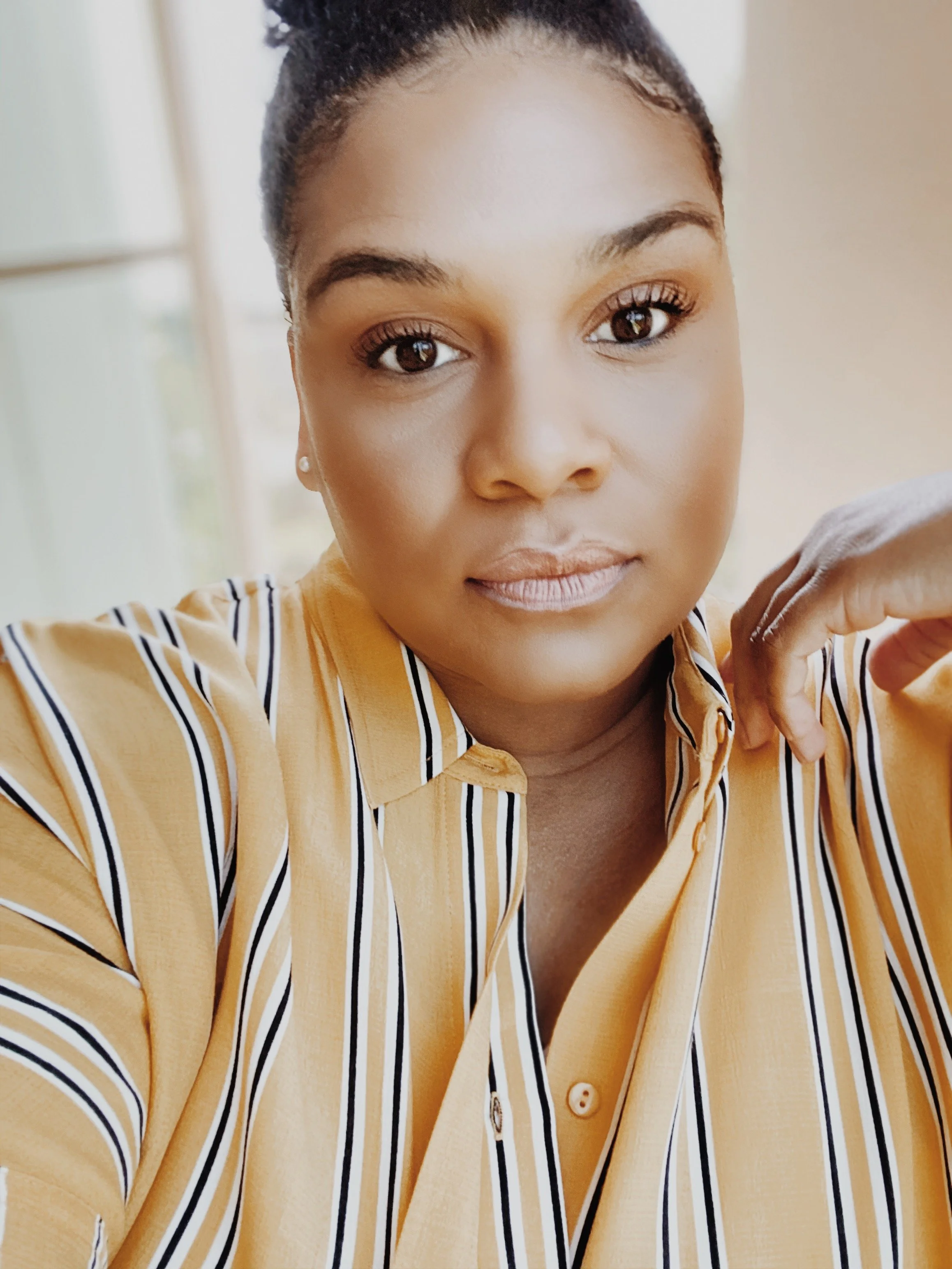 Close-up portrait of a woman with dark skin, wearing a yellow and black striped shirt, looking at the camera with a neutral expression, with a blurred window background.