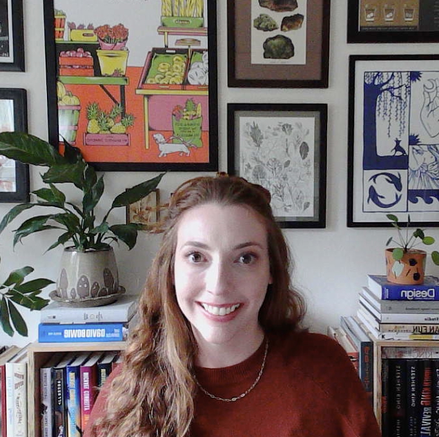A woman with long red hair smiling in a room filled with framed artwork on the wall, a bookshelf, and a large potted plant.