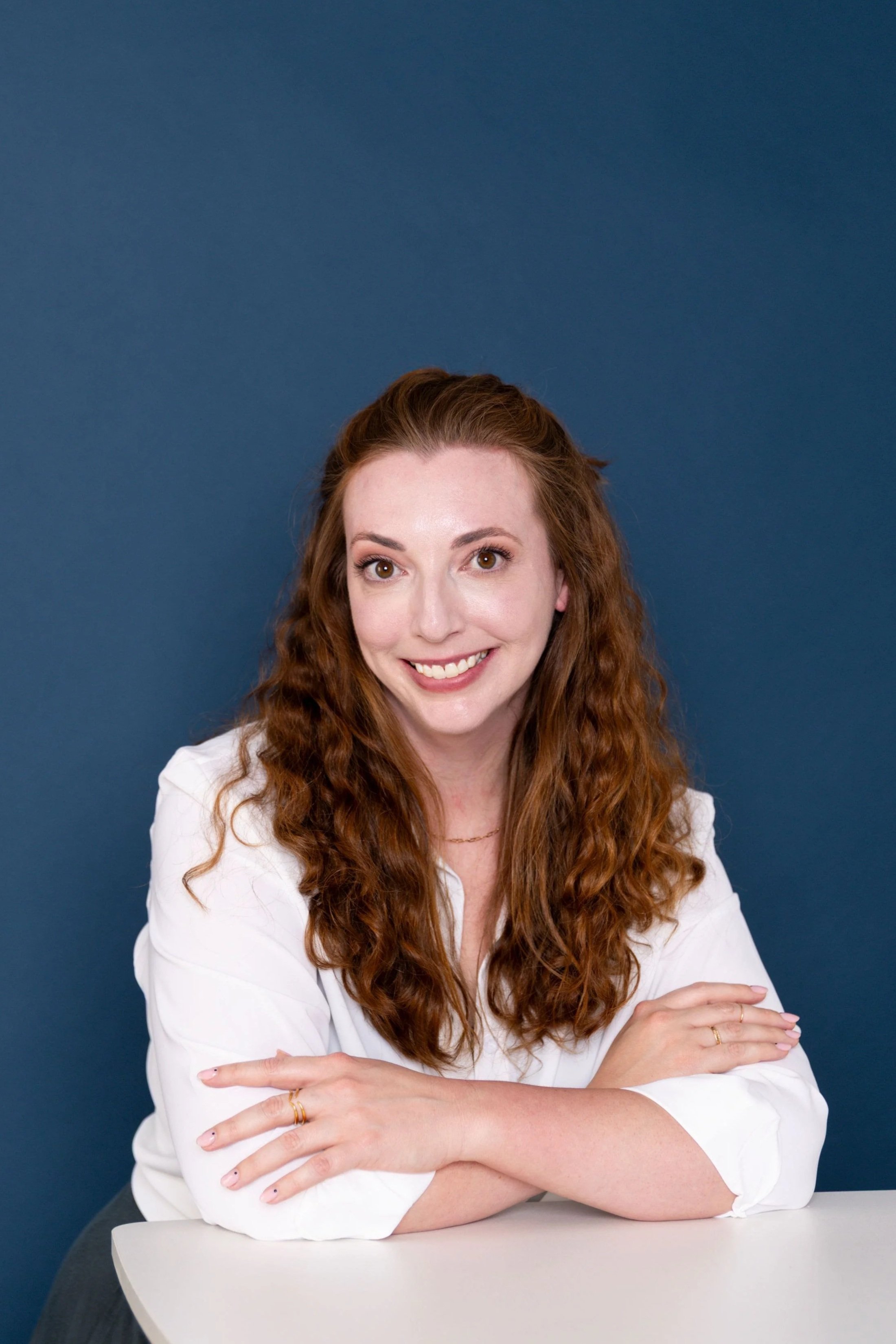 Kat Oldershaw, a woman with long, curly red hair, wearing a white blouse, smiling and sitting with her arms crossed on a white table in front of a blue background.