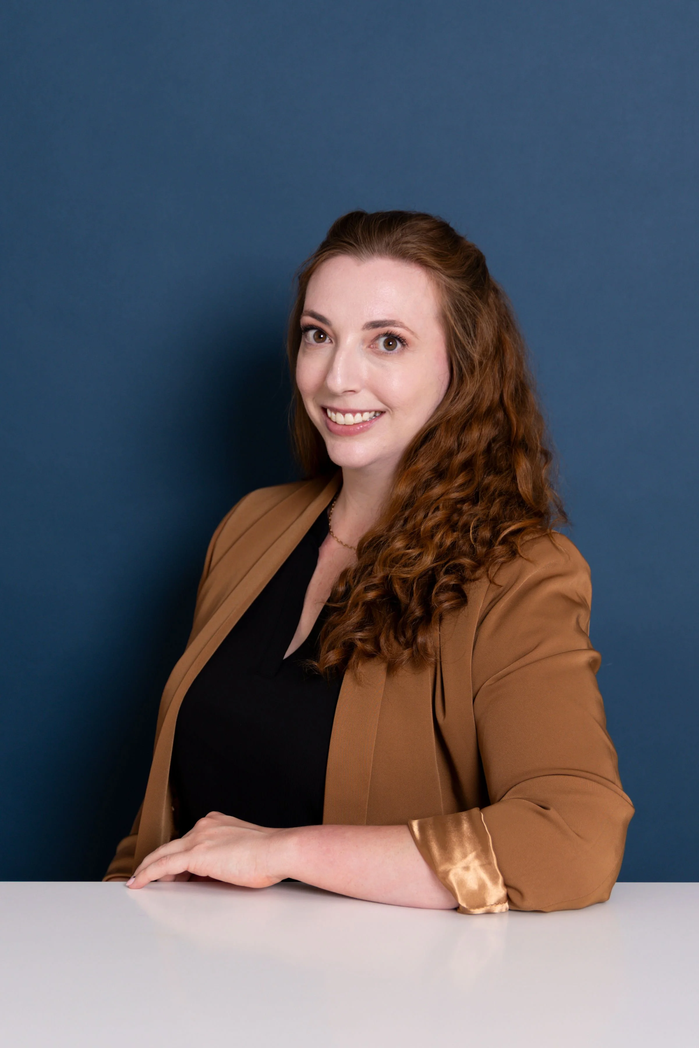 A woman with long, curly red hair, wearing a black top and a brown blazer, sitting at a white desk against a dark blue background, smiling at the camera.