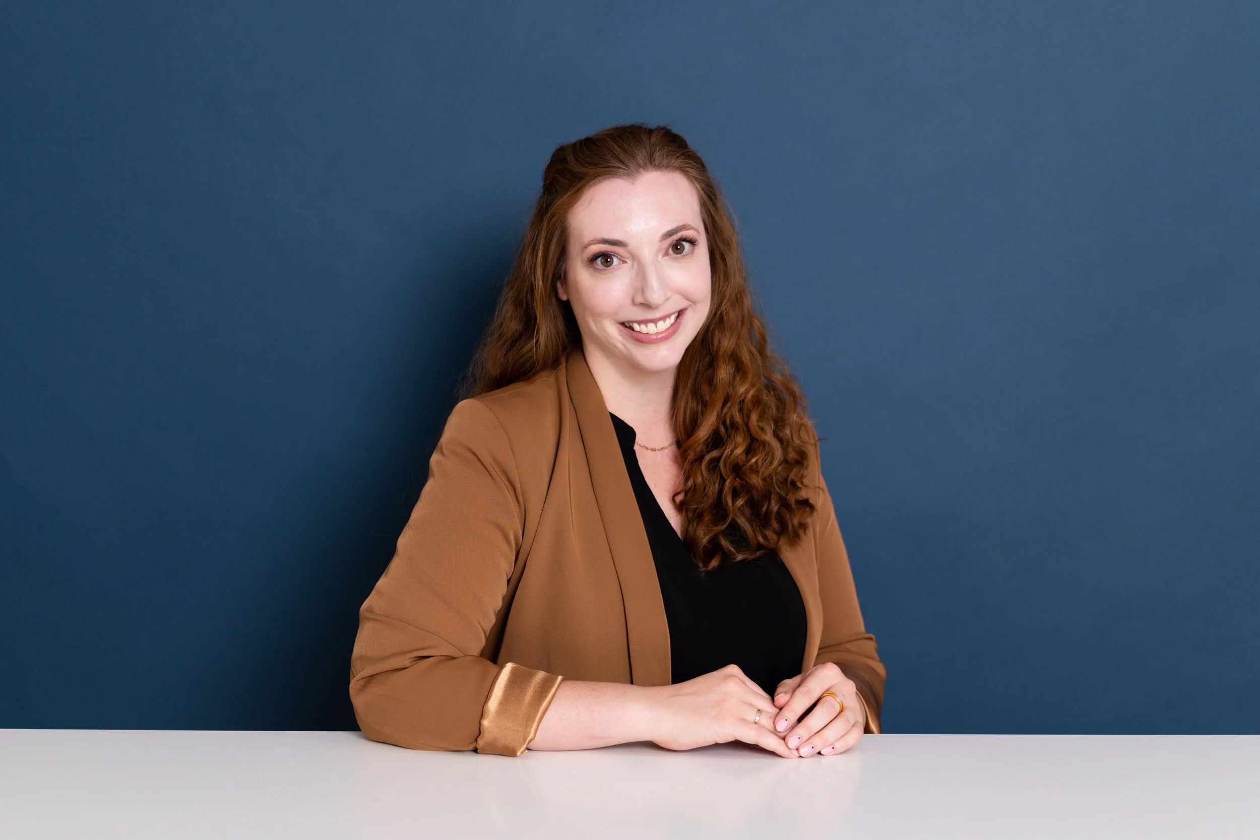A woman with long curly red hair, smiling, wearing a tan blazer and black top, sitting at a table against a blue background.