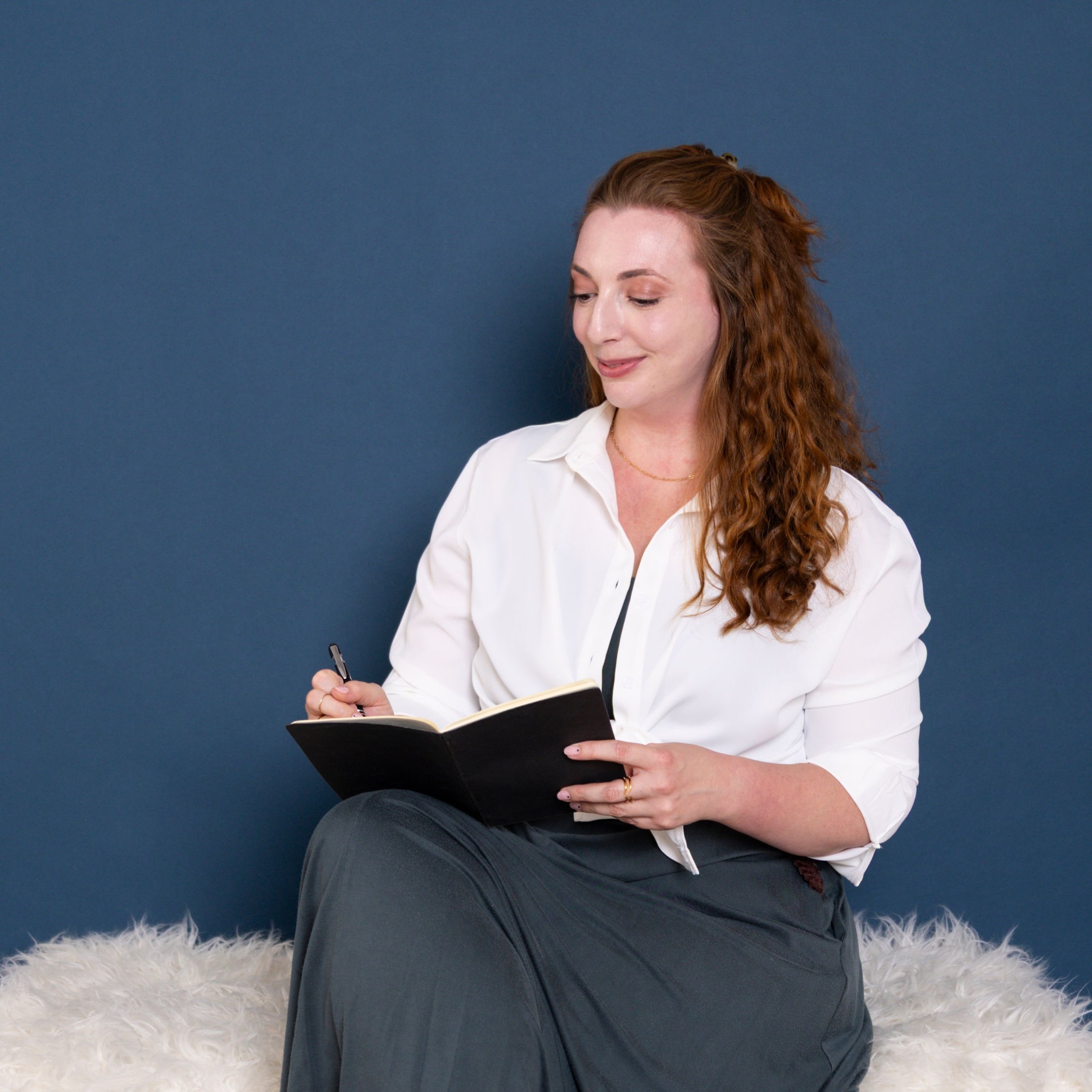 A woman with long, curly red hair, wearing a white blouse and dark pants, sitting on a fluffy white rug against a dark blue wall, holding a black notebook and writing with a pen.