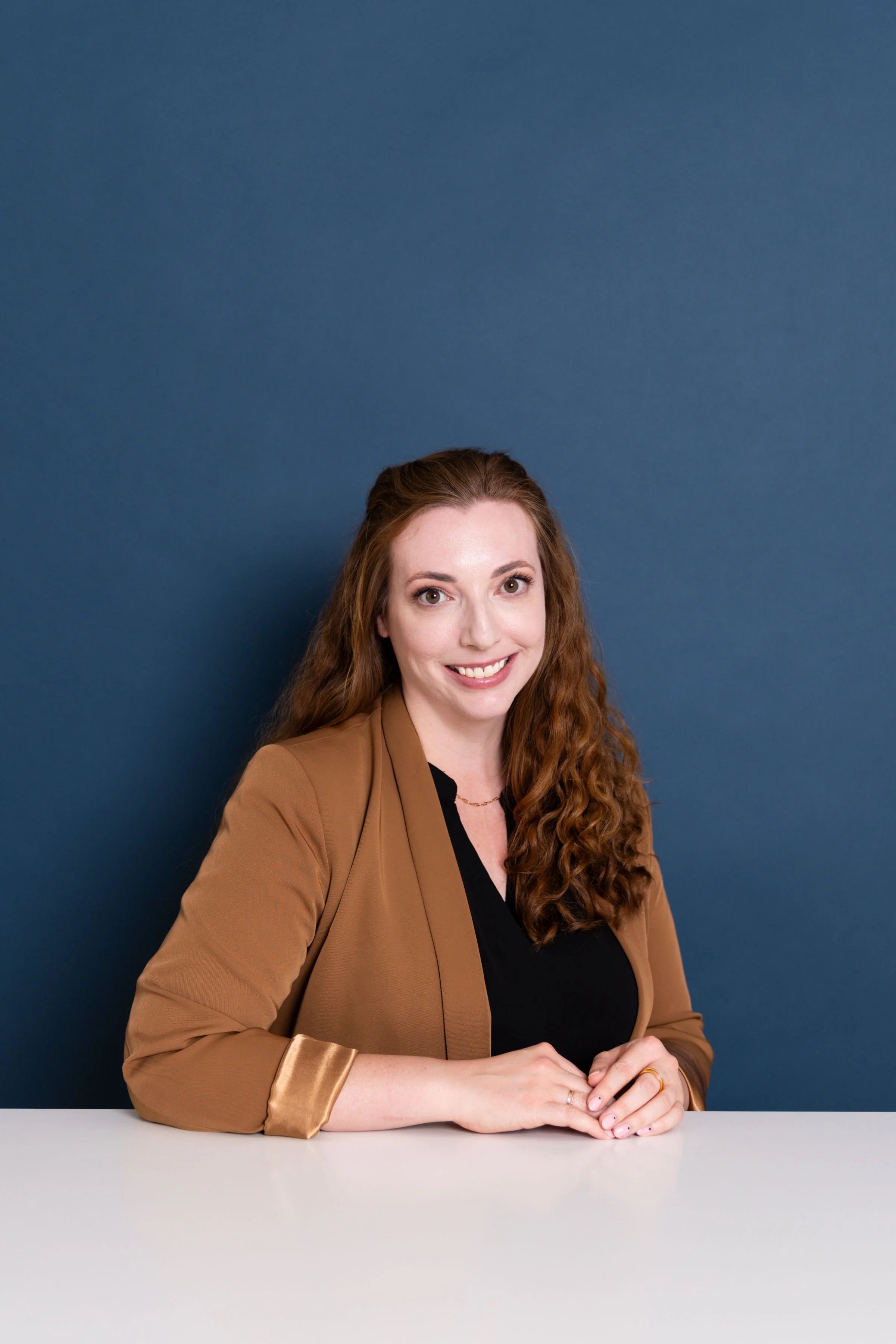 A woman with long curly red hair, smiling, sitting at a table with a white surface, wearing a black top and a tan blazer, against a solid blue background.