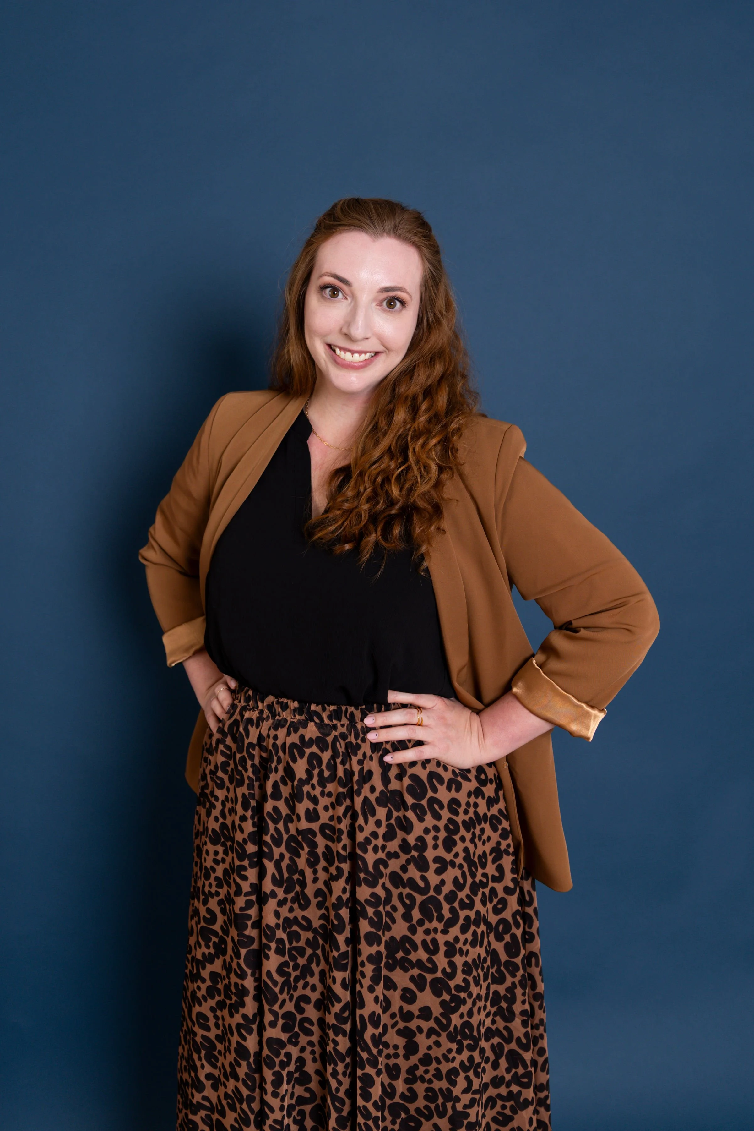 A woman with long curly red hair, wearing a black top, a tan blazer, and leopard print pants, smiling and posing with her hands on her hips against a blue background.