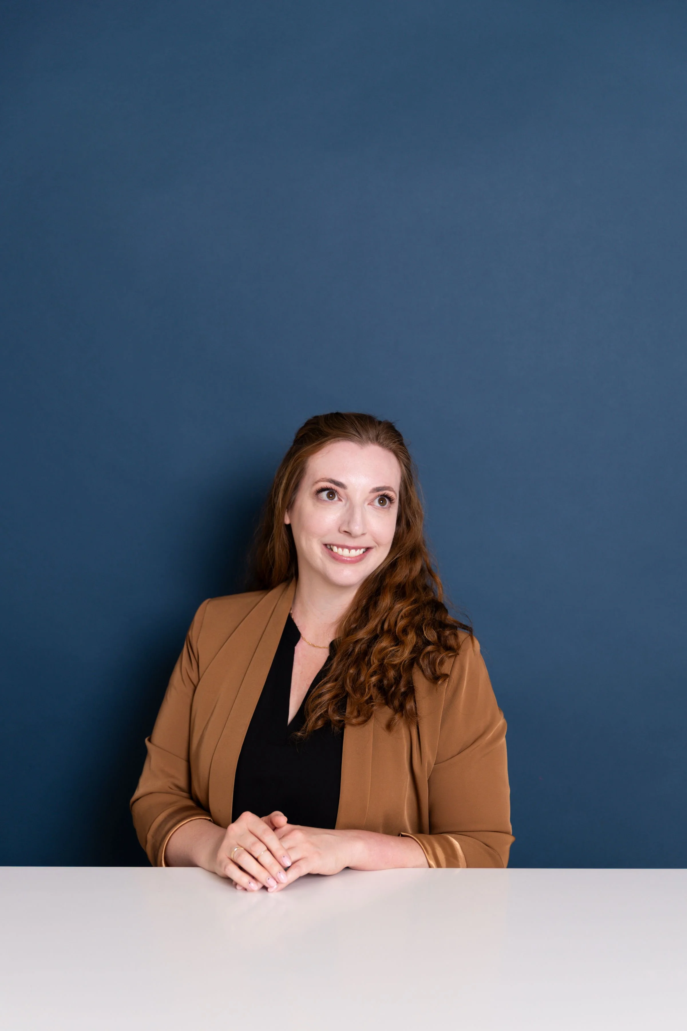 Woman with long curly auburn hair sitting at a white table in front of a blue wall, wearing a black top and brown blazer, smiling and looking slightly to her left.