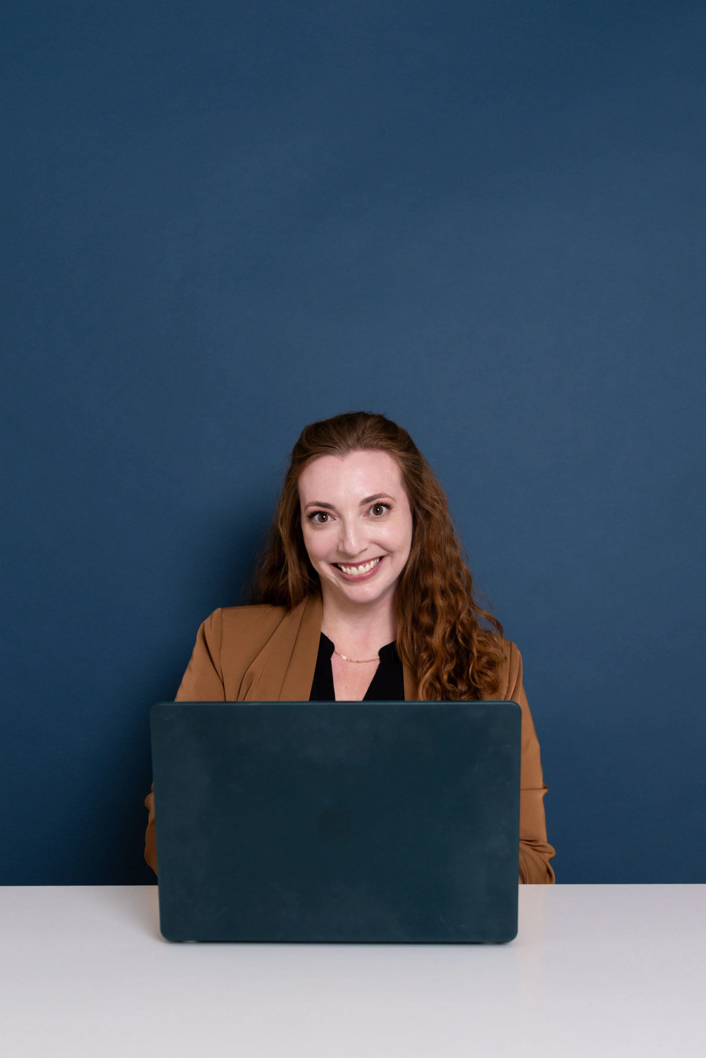 A woman with long curly red hair smiling at a laptop in front of a blue background.