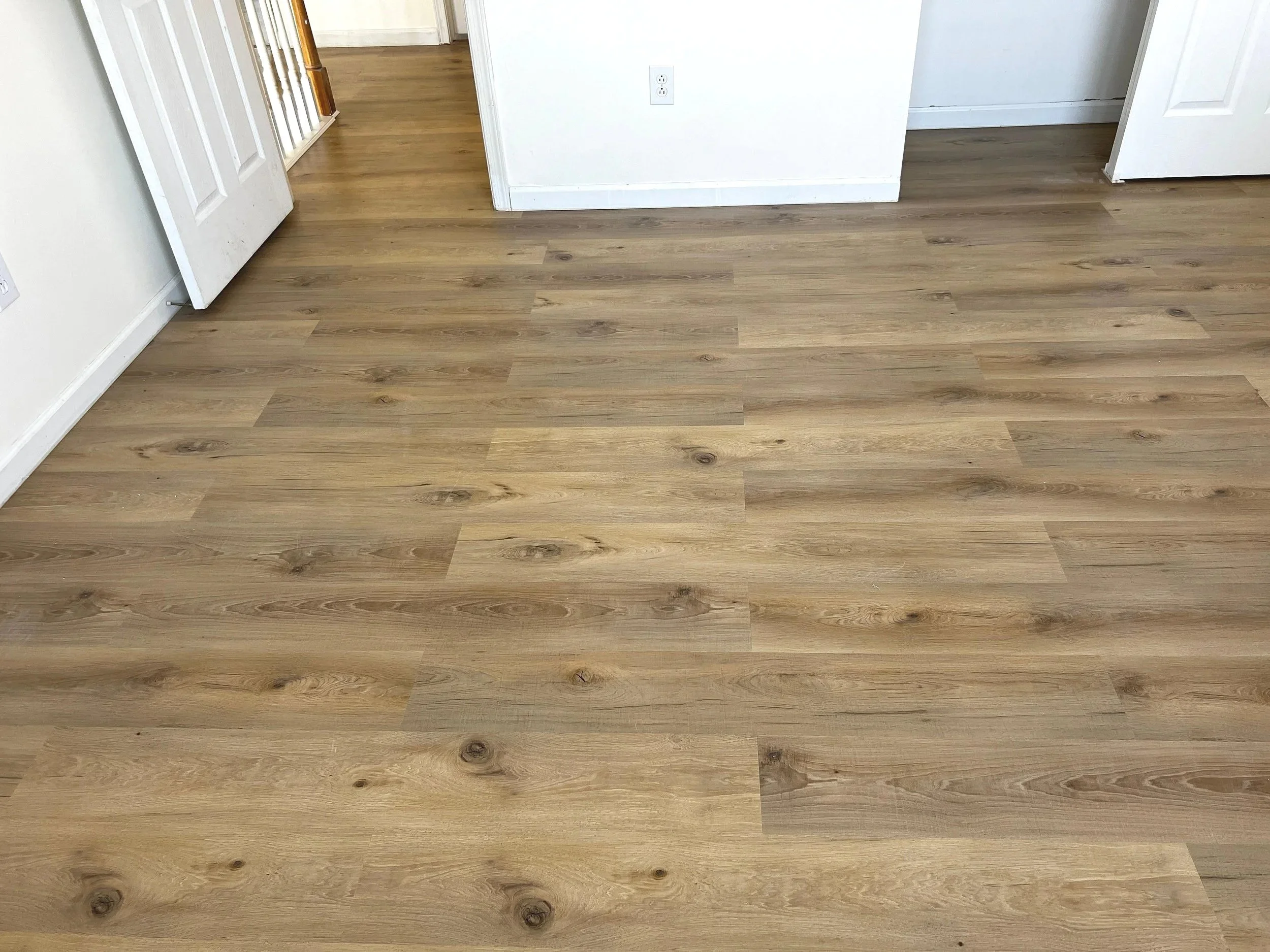 A room with light wood flooring, white walls, and open doorways to other rooms. There is an electrical outlet on the wall.
