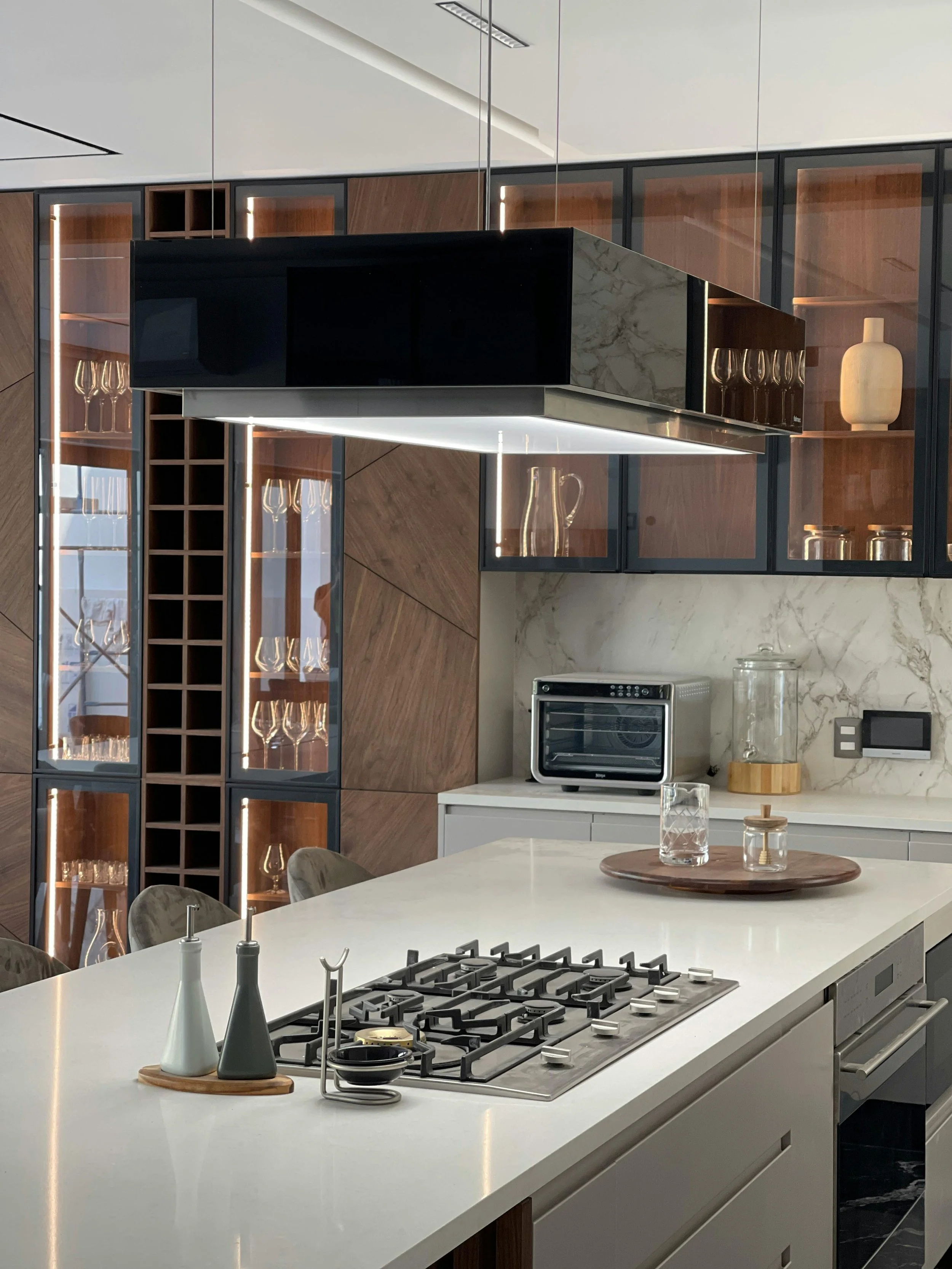 Modern kitchen with island, stove, glass cabinets, decorative items, and a black overhead vent hood.