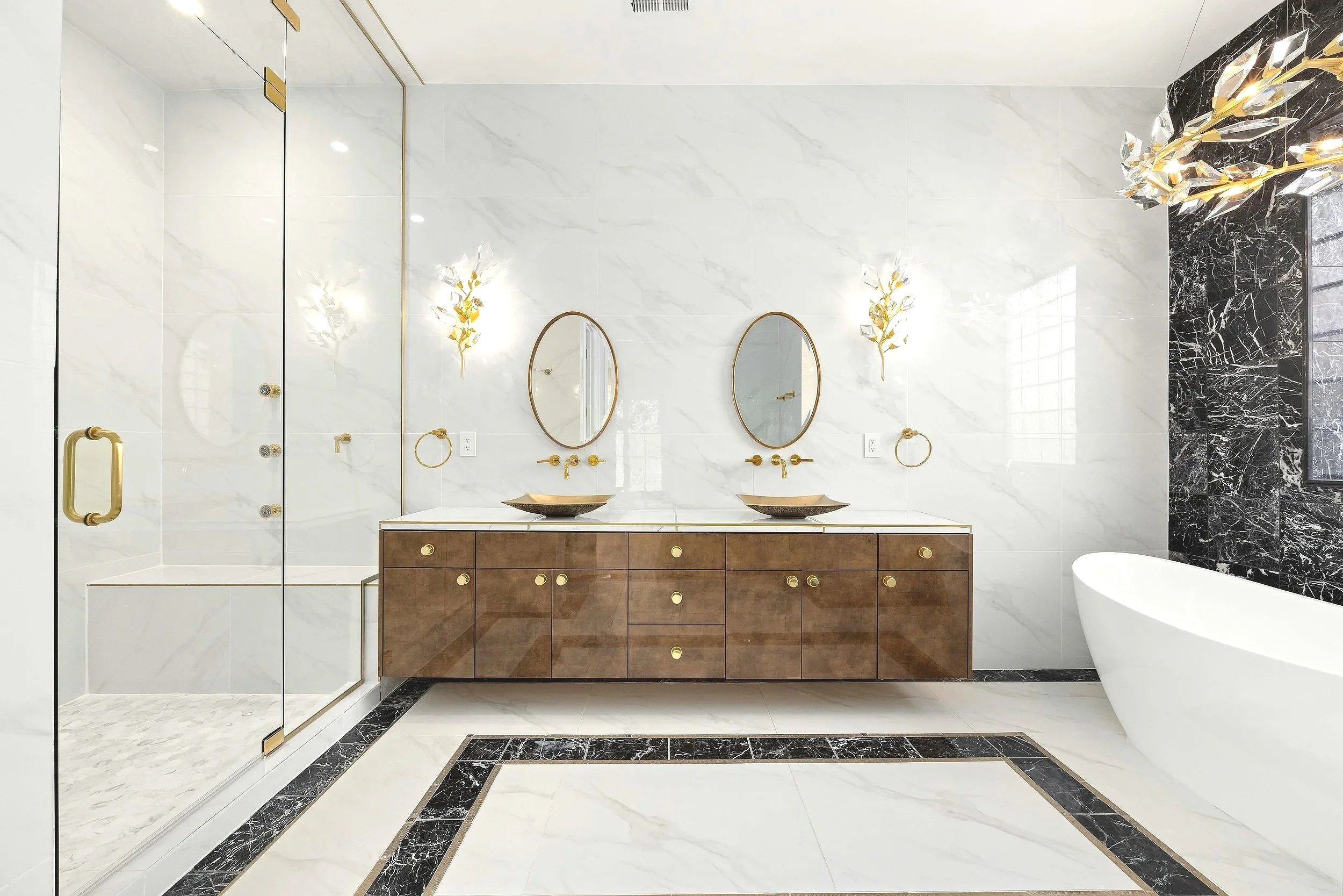 Modern bathroom with a double vanity with two sinks, oval mirrors, wall-mounted faucets, gold accents, a glass-enclosed shower, and a freestanding bathtub, with marble walls and black marble accents.