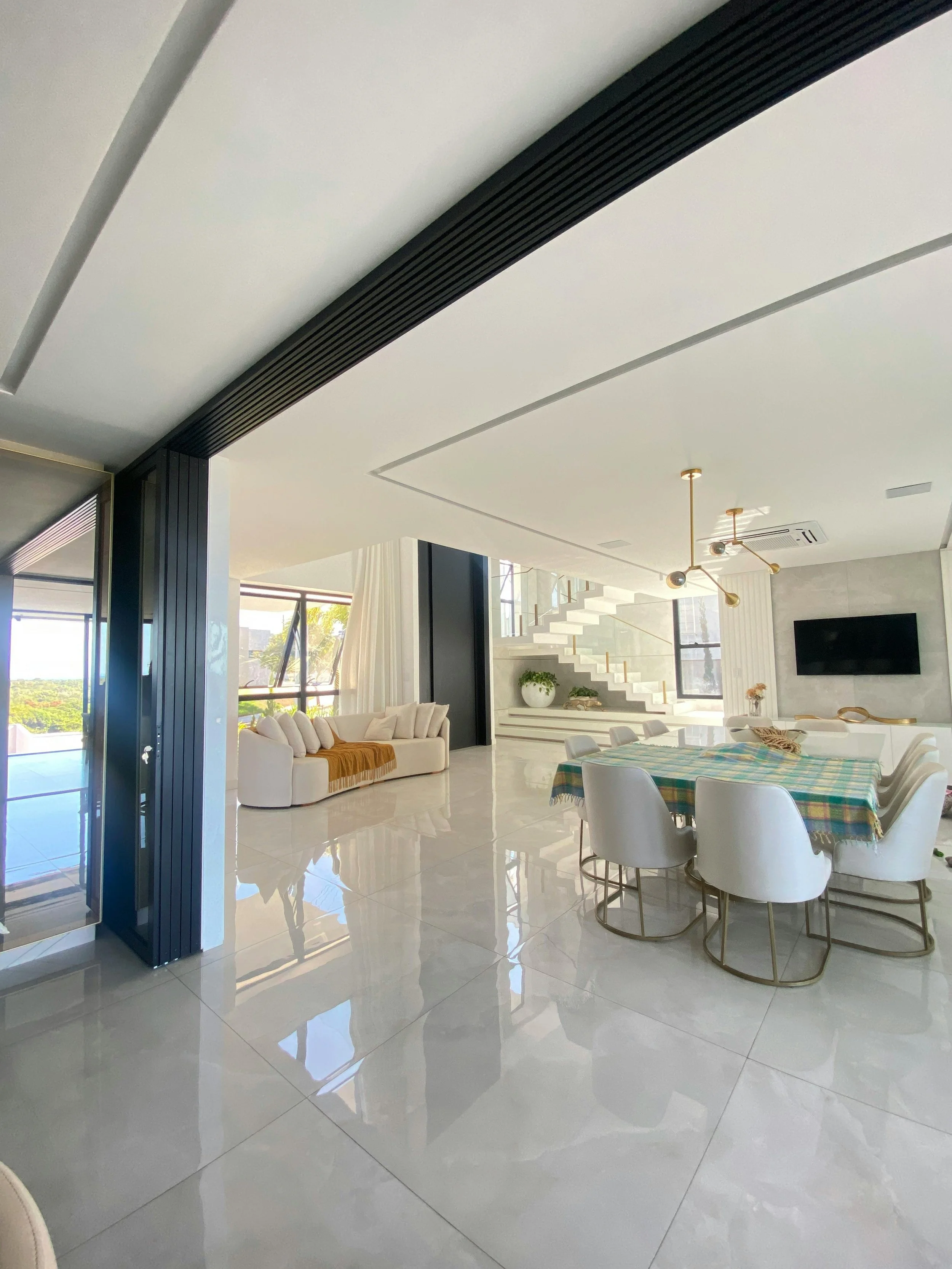 Bright, modern living room with white tiled floors, a dining table with a plaid tablecloth, white chairs, a white sofa with cushions, large windows with curtains, and a staircase with a glass railing.