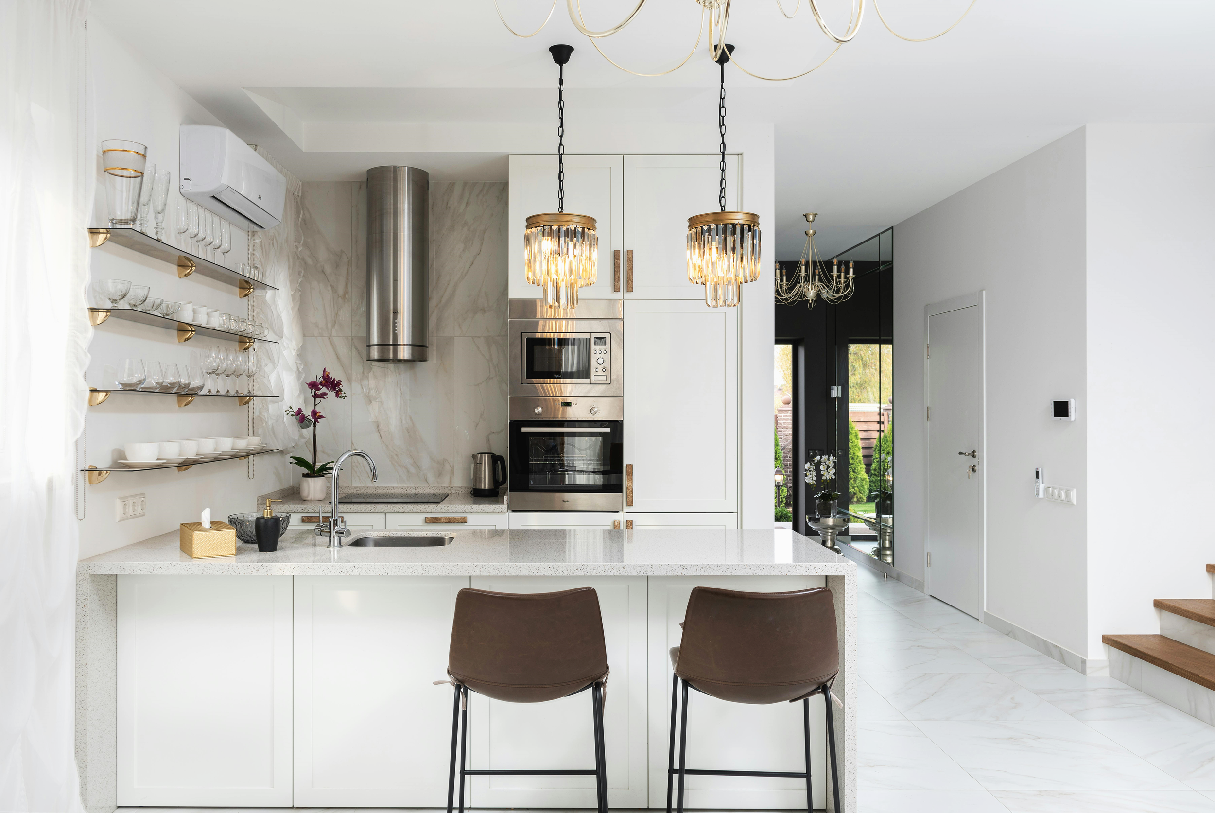 Modern kitchen with white cabinets, marble backsplash, and marble countertops, featuring two hanging chandeliers above the island, two brown chairs at the island, and open shelving with glassware and dishes on the left.