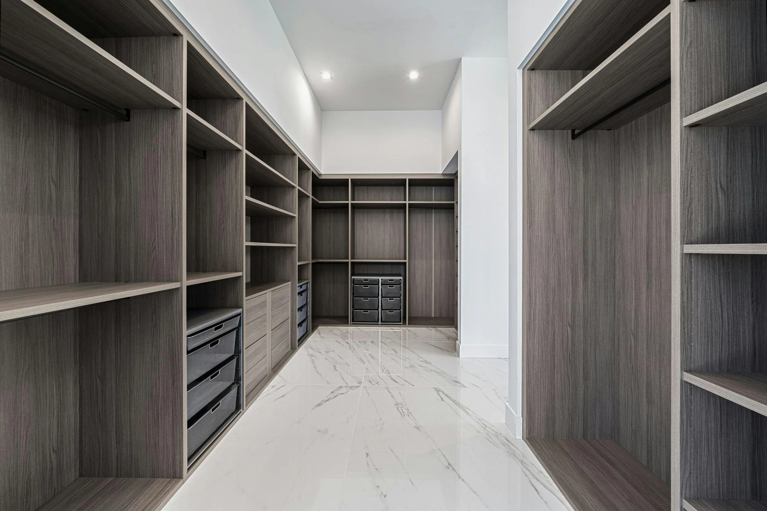 Empty walk-in closet with dark wood shelves and drawers, white walls, and marble tile floor.