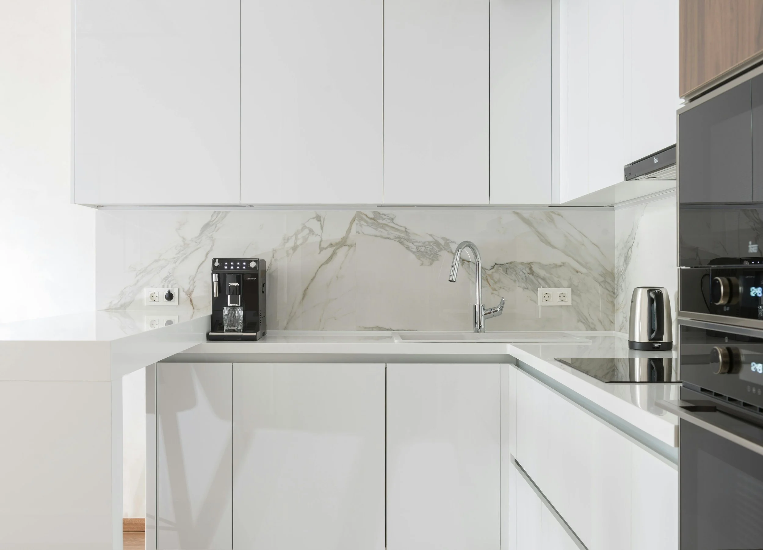 Modern kitchen with white cabinets, marble backsplash, and small appliances including a coffee machine, electric kettle, and a built-in oven.