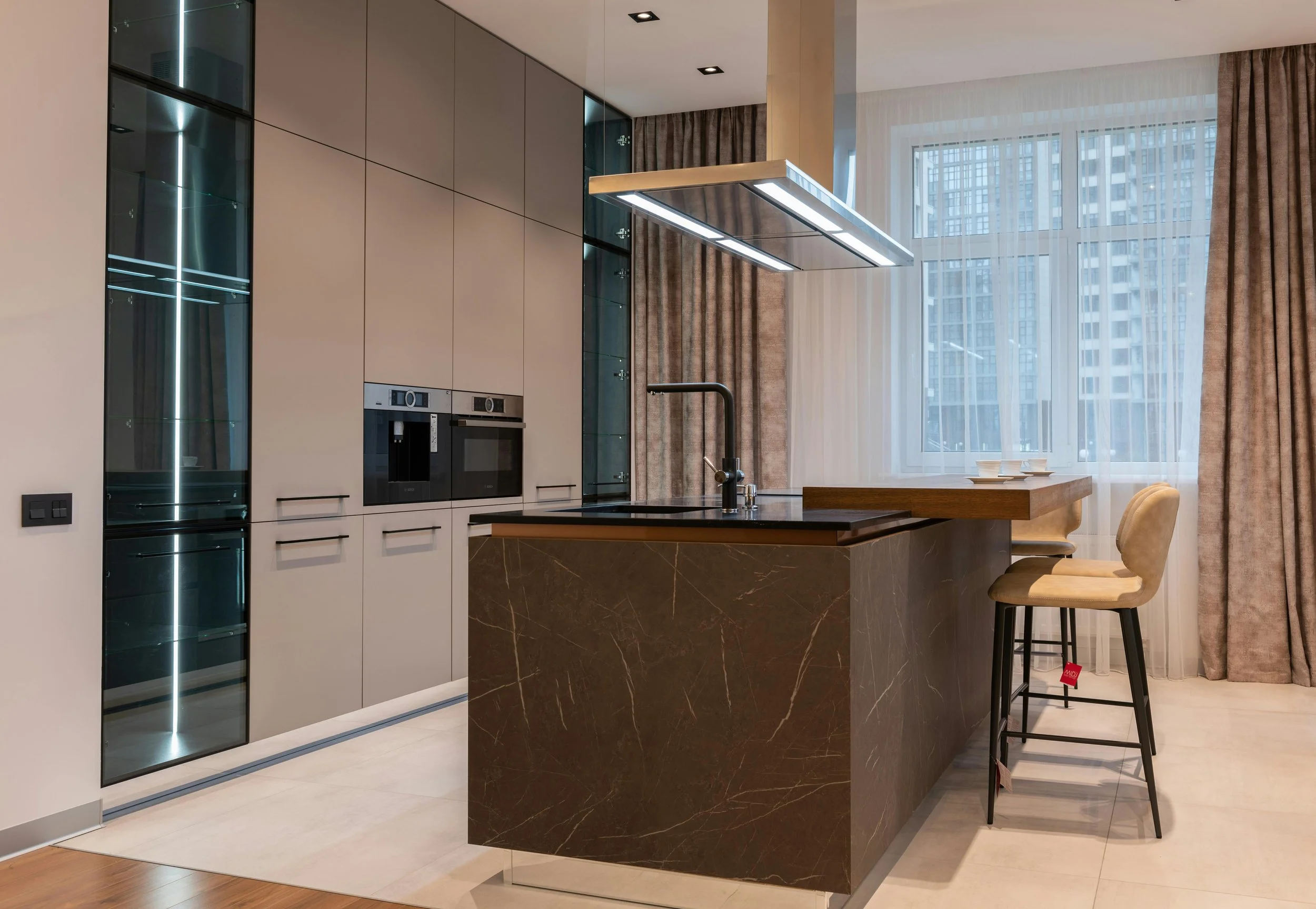 Modern kitchen with black marble island, beige cabinetry, built-in microwave and oven, glass display cabinet, and large window with city view.