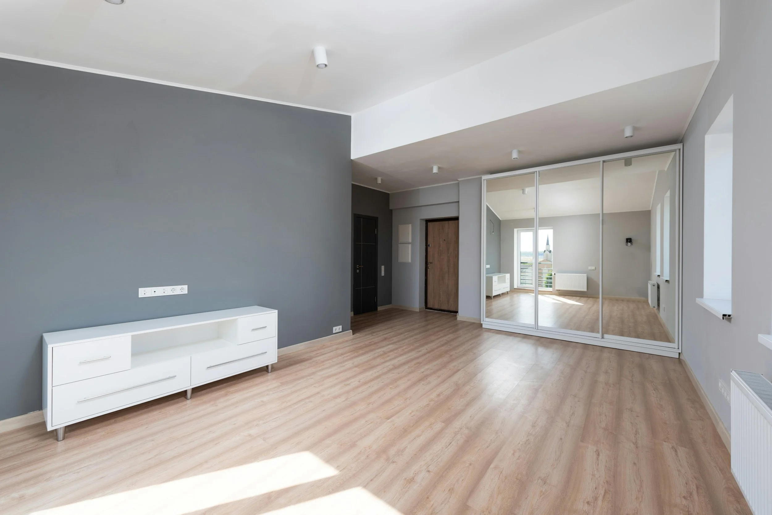 Empty living room with light wood floors, gray and white walls, a large mirror closet, and a door leading to a balcony.