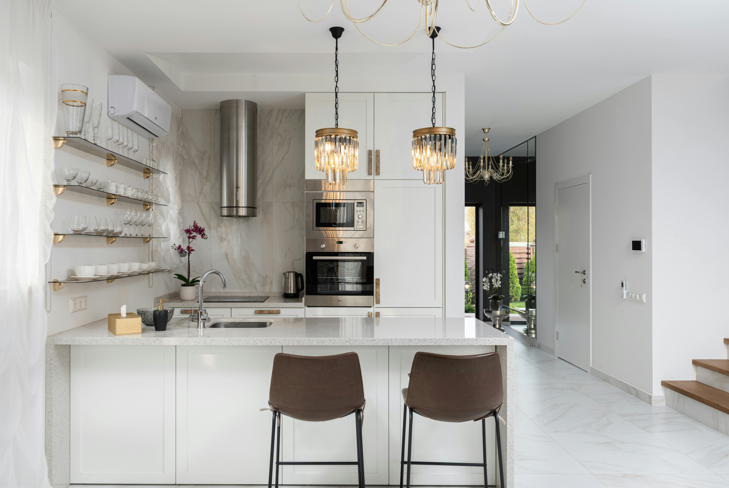 Modern white kitchen with island, brown chairs, and decorative lighting fixtures. Open view to patio with greenery outside.
