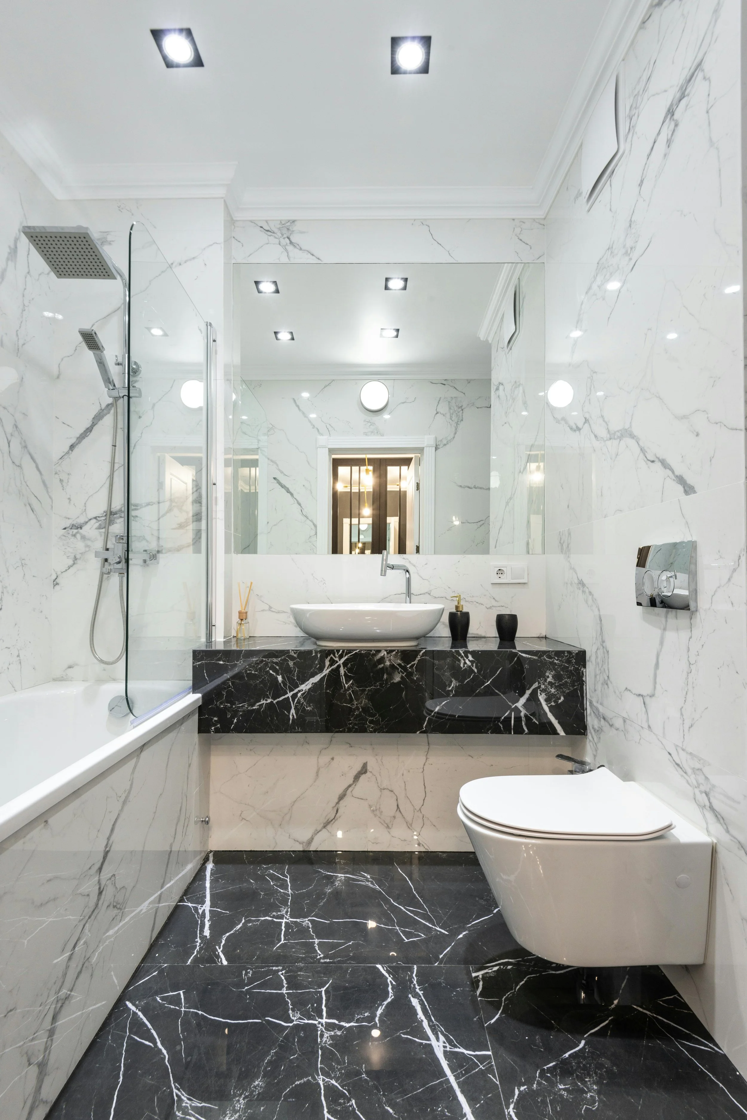 Modern bathroom with black and white marble walls and floor, a bathtub with a glass shower screen, a large mirror, a vessel sink on a black marble vanity, black vases, a wall-mounted toilet, and ceiling lights.