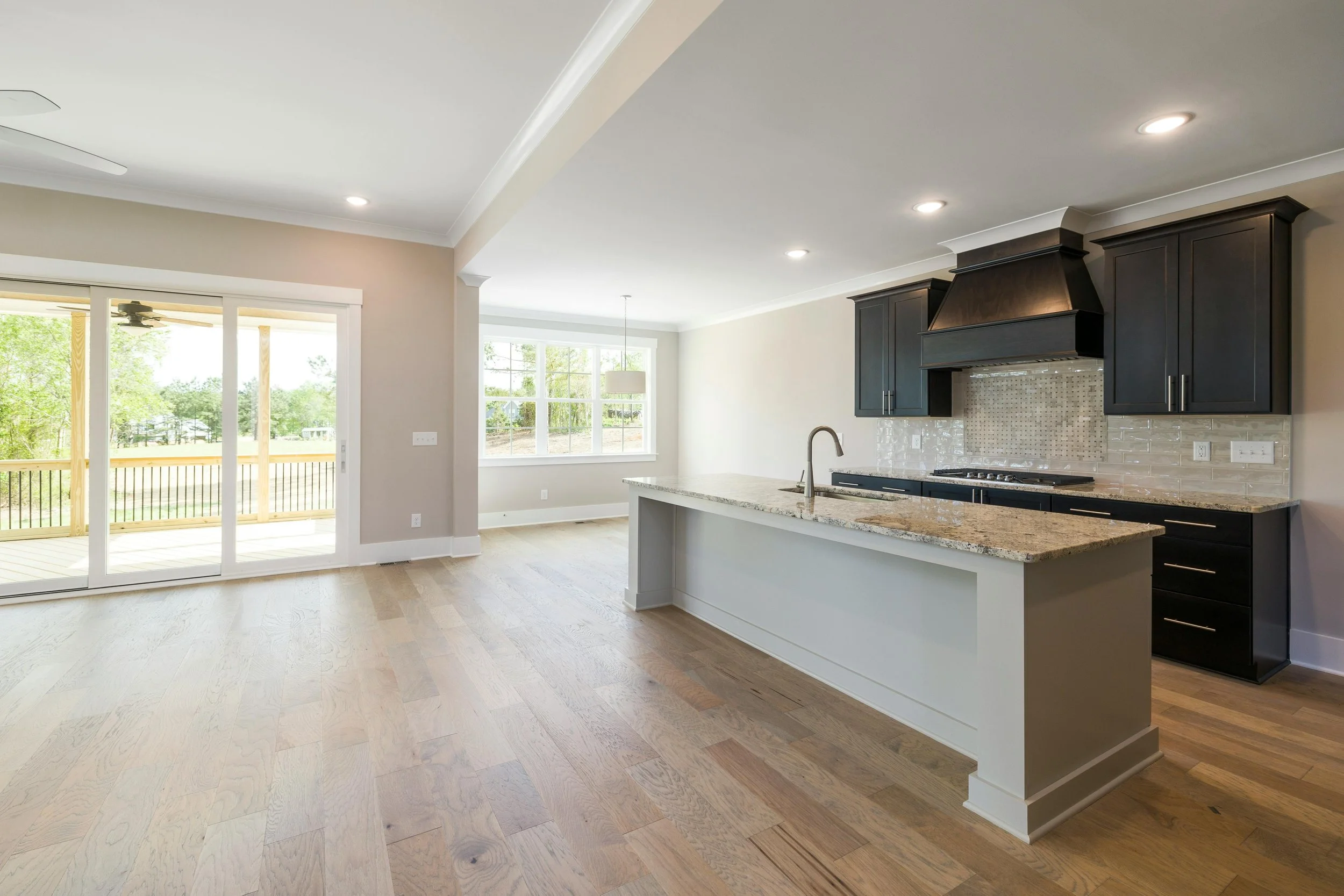 Open-concept kitchen with black cabinets, granite countertops, a white island, and a backsplash in a modern home, with large windows and sliding glass door leading to a porch.
