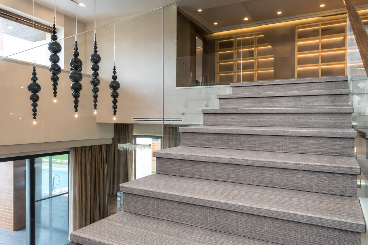 Modern interior staircase with gray carpeted steps, glass railing, and black beaded pendant lights, overlooking a living area with large windows and curtains.