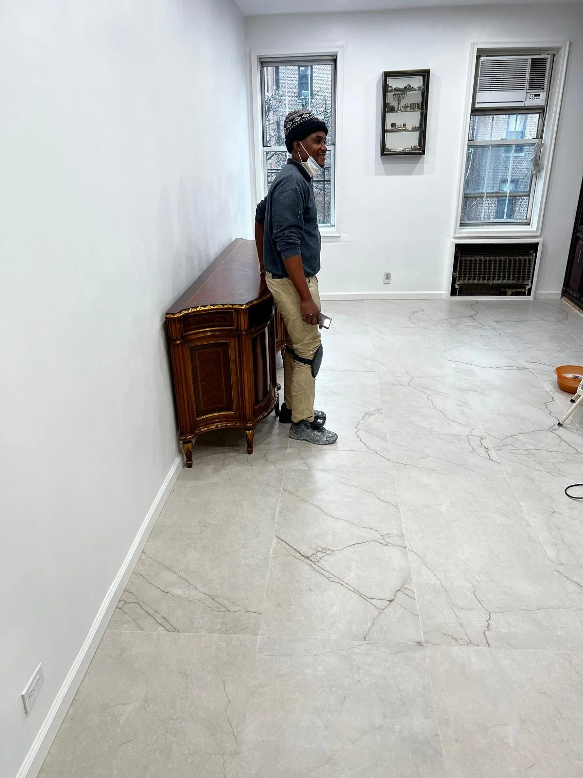 A man in work clothes stands in a bright, empty room with white walls, marble floors, and large windows with city views. He is smiling and leaning against an ornate wooden sideboard.