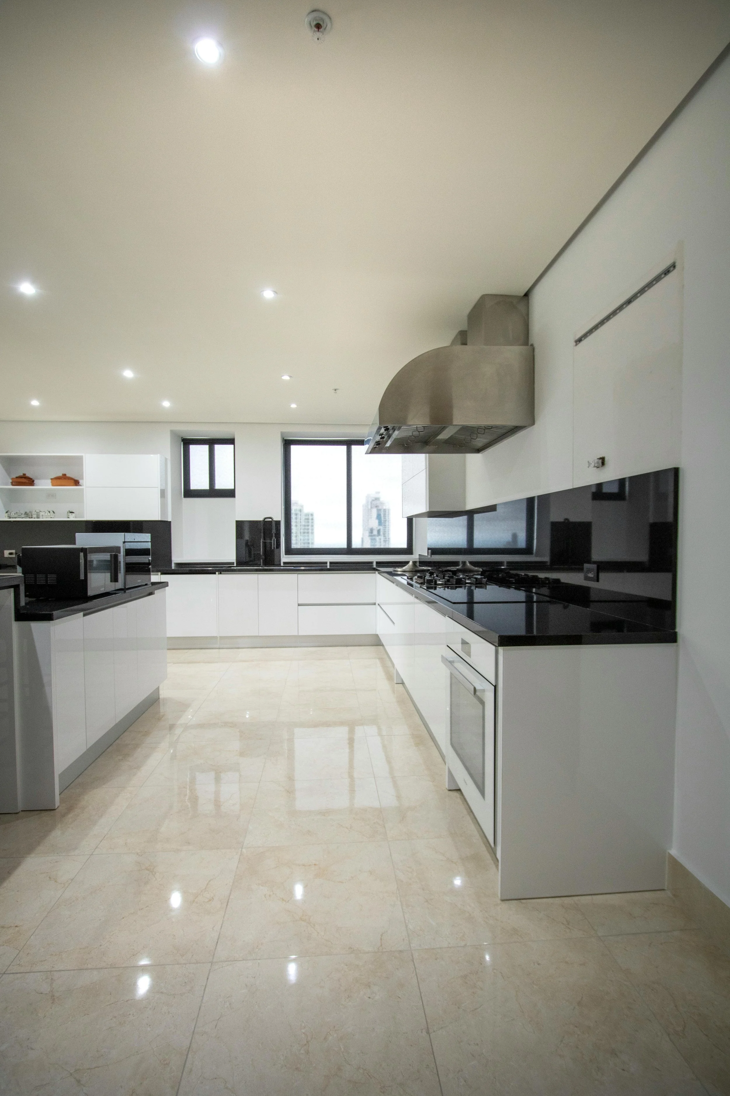Modern kitchen with white cabinets, black countertops, stainless steel range hood, and beige tiled floor, with multiple windows providing natural light.