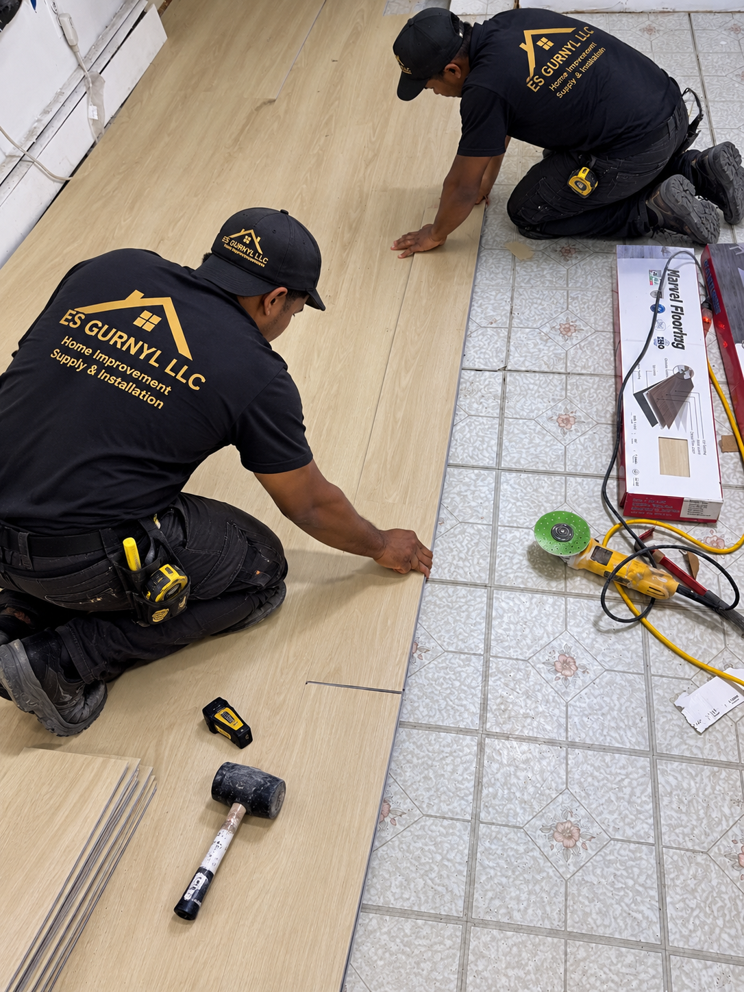 Two workers from ES Gurny LLC installing a wood or laminate flooring over a tiled floor, with tools such as a hammer, measuring tape, and a power saw nearby.
