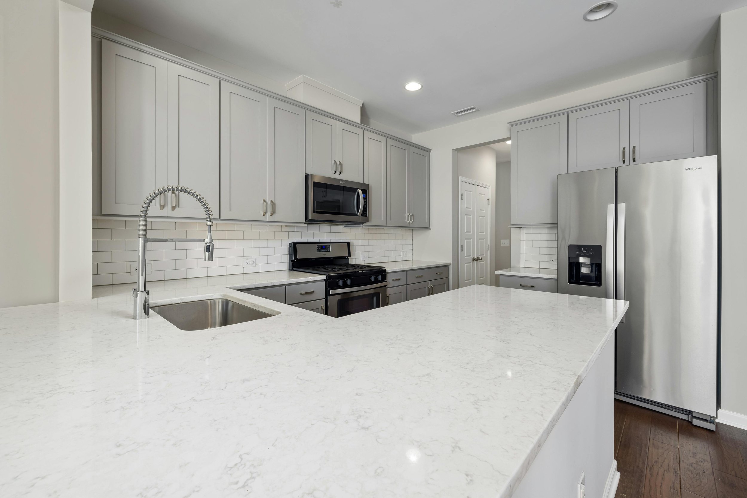 A modern kitchen with white quartz countertops, gray cabinets, stainless steel appliances including a microwave, stove, and refrigerator, white subway tile backsplash, and a sink with a high-arc faucet.