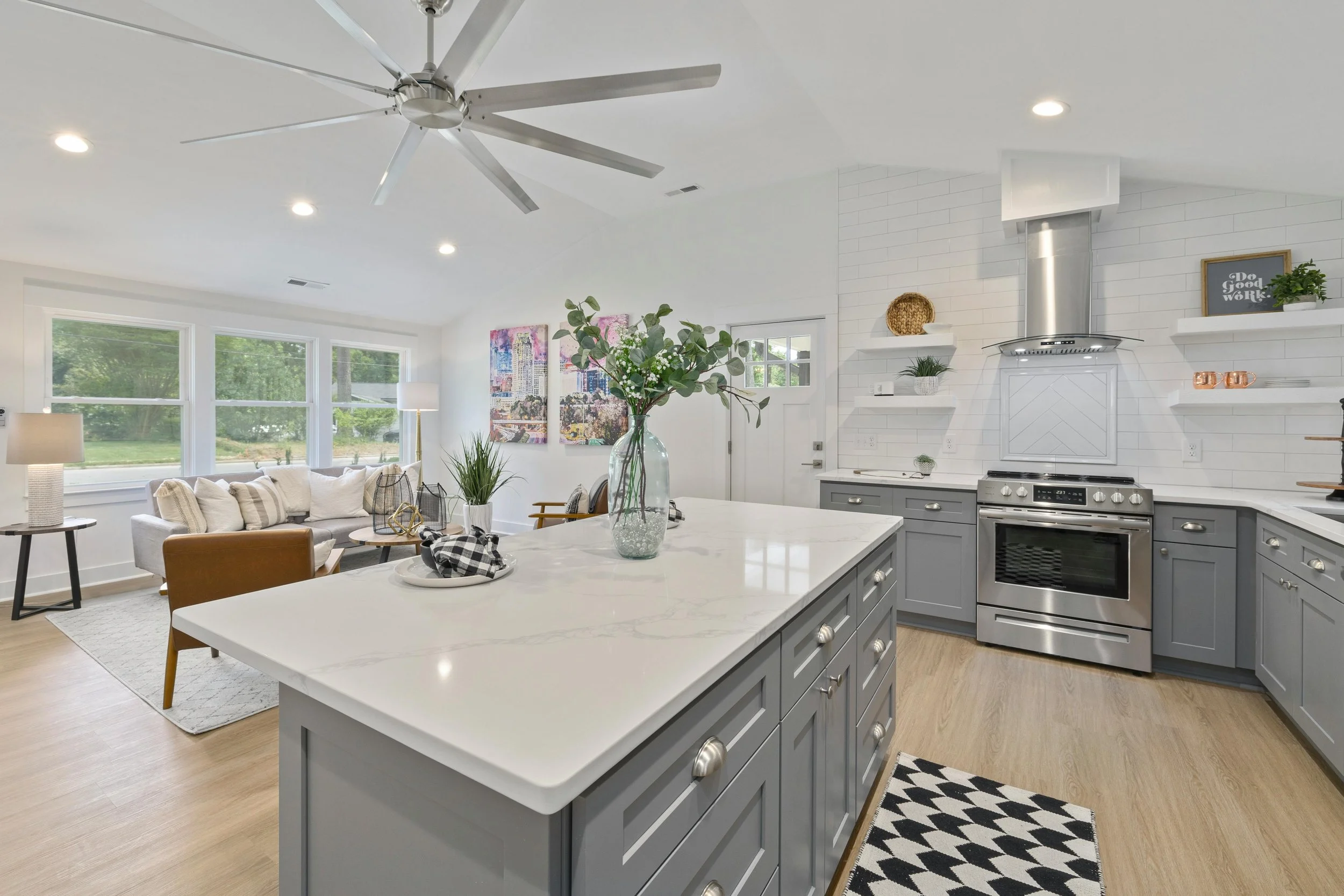 Open-concept kitchen and living room with large island, gray cabinets, stainless steel appliances, white brick backsplash, and cozy seating area with large windows, houseplants, and artwork.