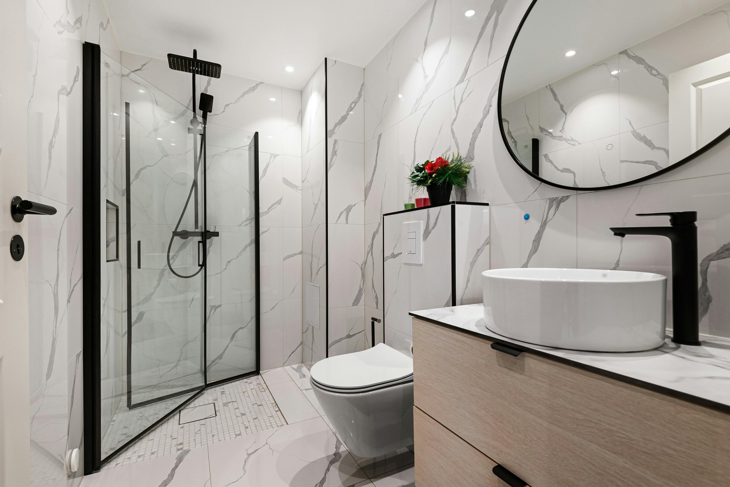 Modern bathroom with white marble tiles, a glass shower enclosure with black fixtures, a toilet, a wooden vanity with a vessel sink, a round wall mirror, and a small shelf with a plant.