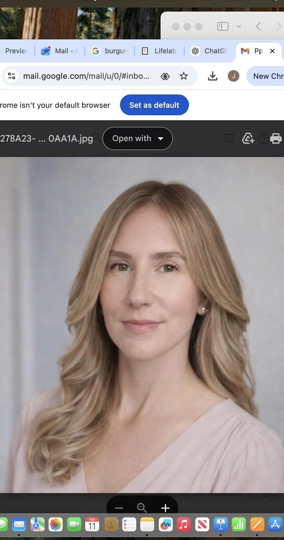 A young woman with light brown, wavy hair, wearing a white button-up shirt and a small necklace, standing in front of a plain, off-white background.
