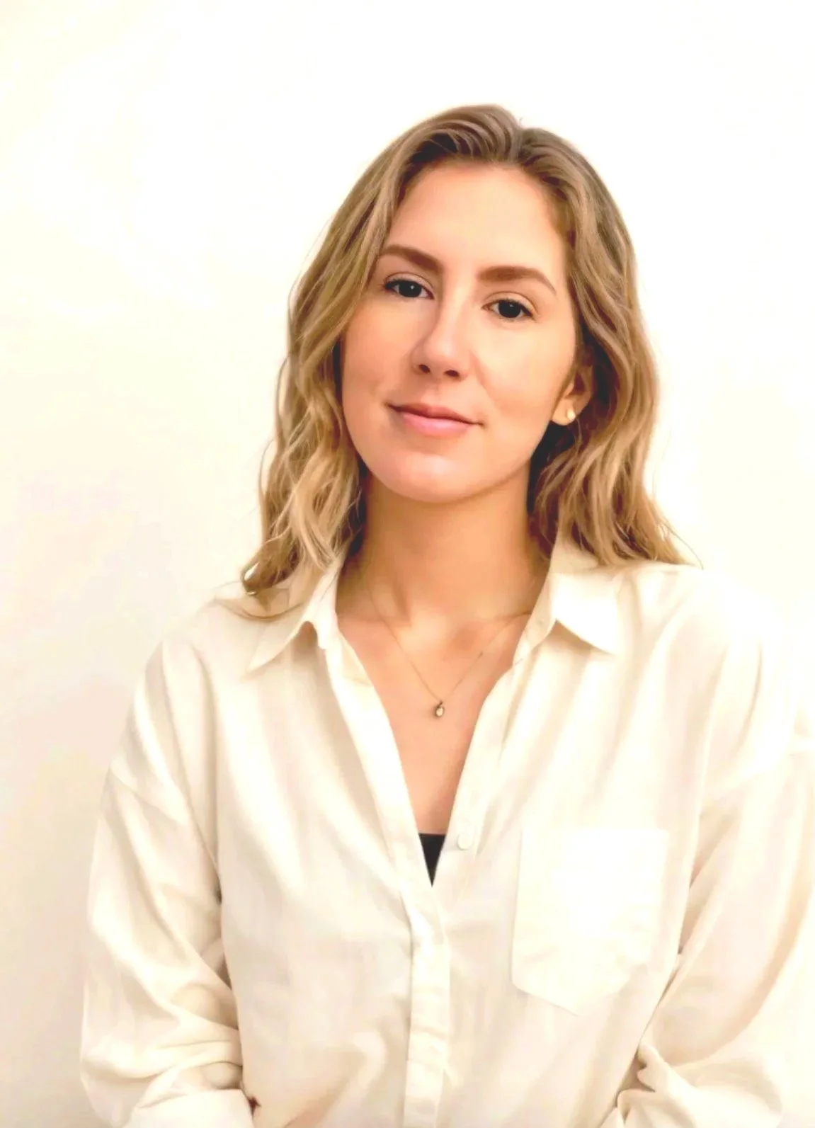 A young woman with light brown, wavy hair, wearing a white button-up shirt and a small necklace, standing in front of a plain, off-white background.