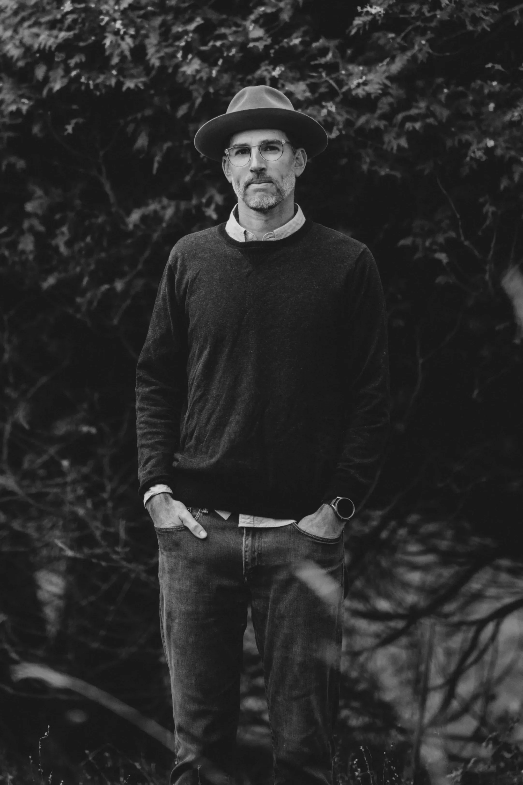 A black-and-white photo of a man standing outdoors in front of trees, wearing a fedora hat, glasses, a sweater over a collared shirt, jeans, and a watch, with hands in his pockets.