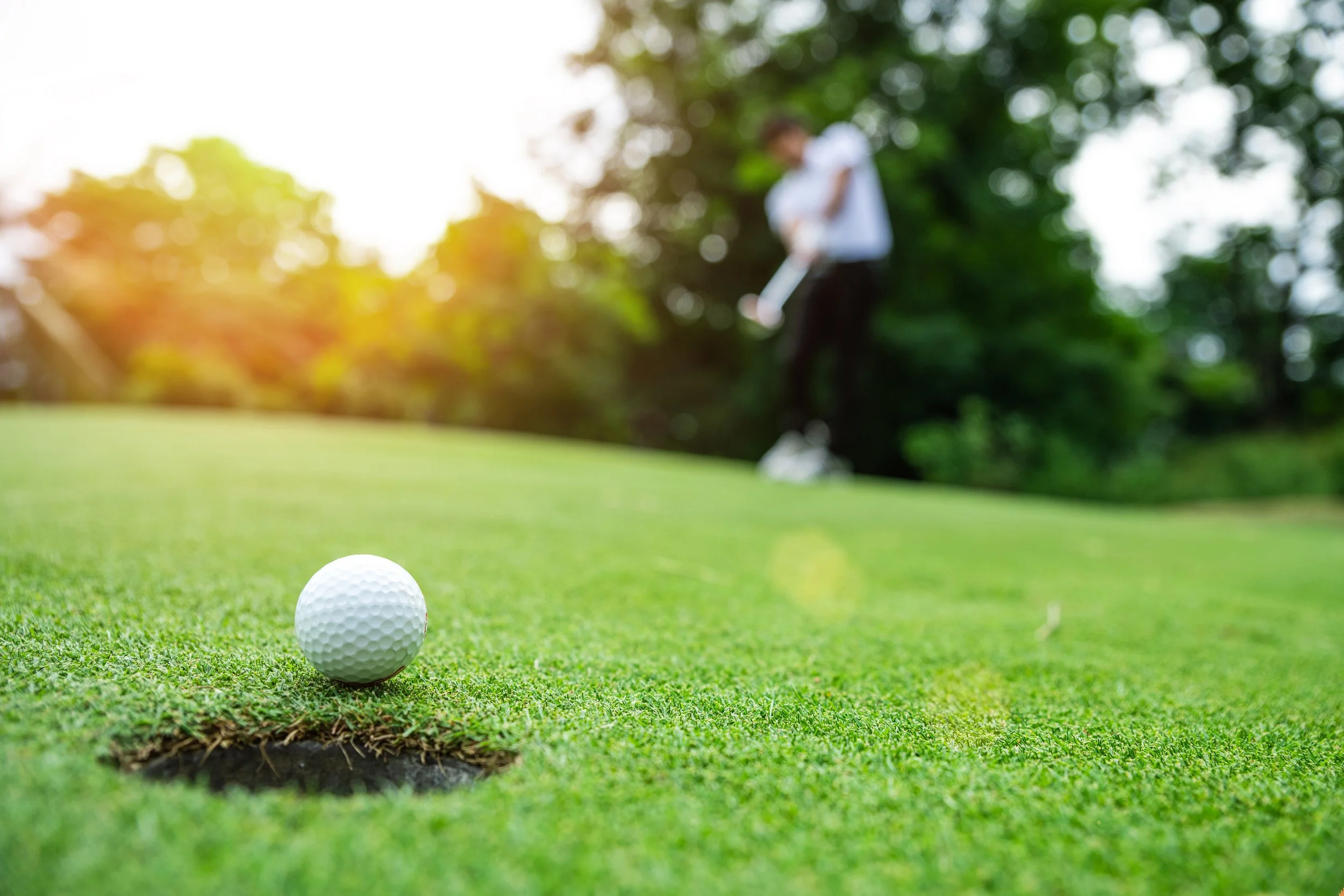 Close-up of a white golf ball on the green grass near a hole on a golf course, with a blurred golfer in the background preparing to hit the ball.