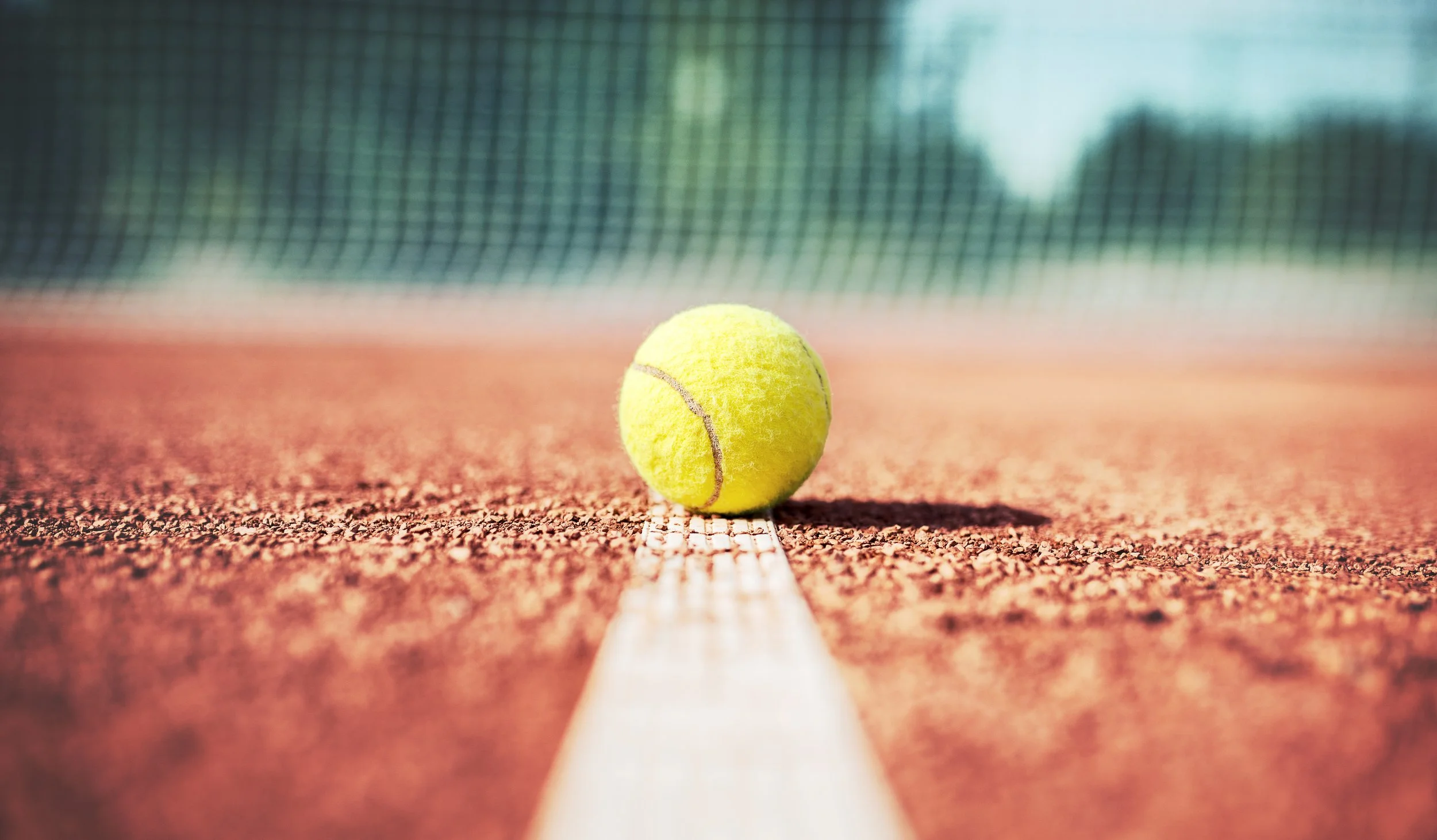 Close-up of a yellow tennis ball resting on the white baseline of a clay tennis court, with a tennis net in the background.