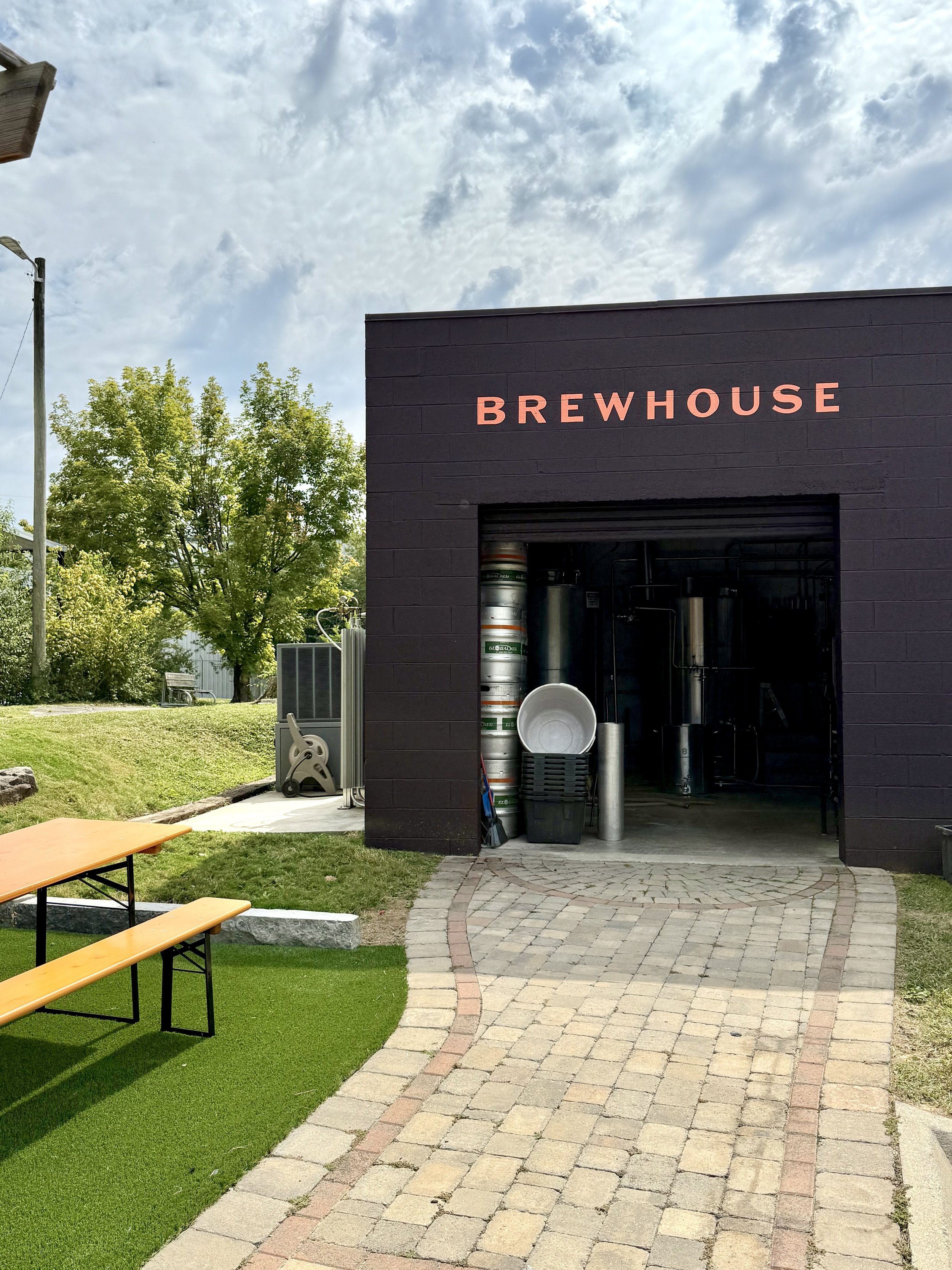 Exterior of a brewpub with a black building labeled 'BREW HOUSE', a paved pathway, outdoor seating with a bench and table, and brewing equipment inside visible through an open garage-style door.
