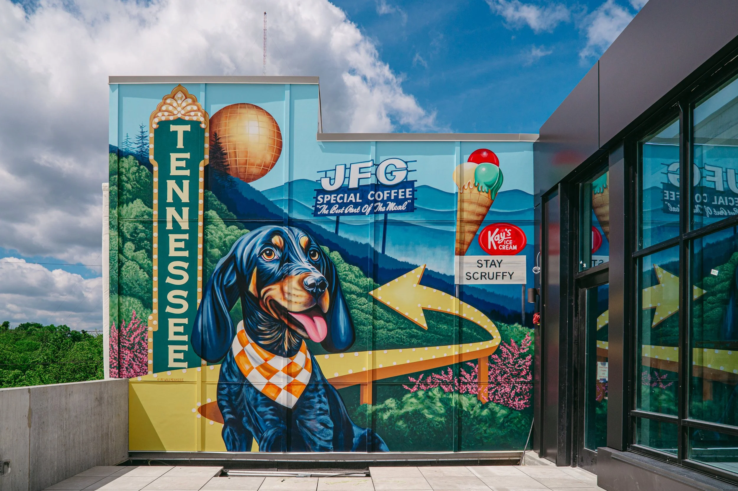 Colorful mural of a black and tan dog wearing a checkered bandana, with a mountain landscape, trees, and pink flowers in the background, and a large ice cream cone on the right, adjacent to a building with glass windows.