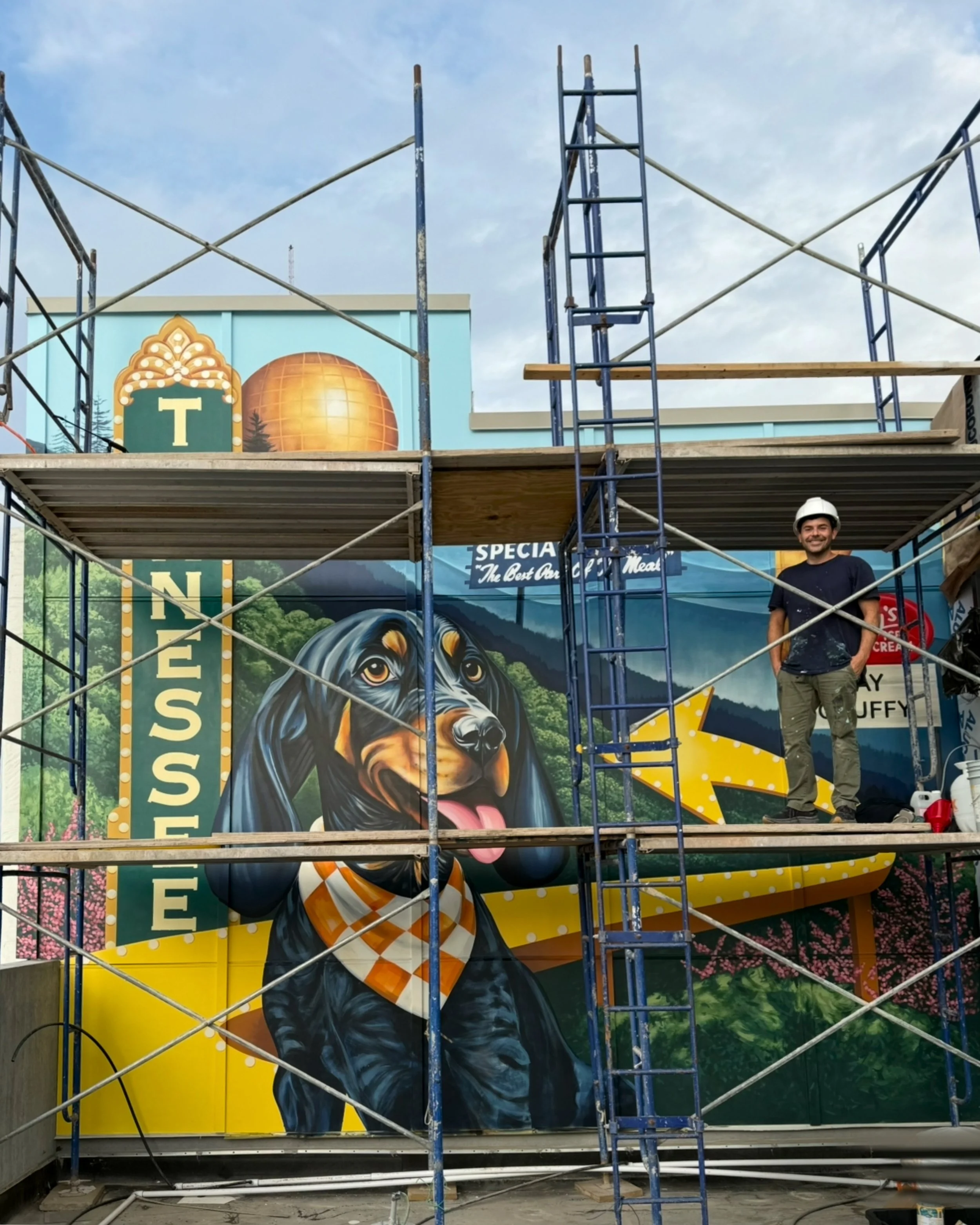 A man standing on construction scaffolding in front of a colorful mural of a black dog with a checkered bandana, with the words "Business" and "Special" visible, and a yellow arrow. Part of a sign with a large letter "T" and a decorative border, as well as a landscape background, are also visible.