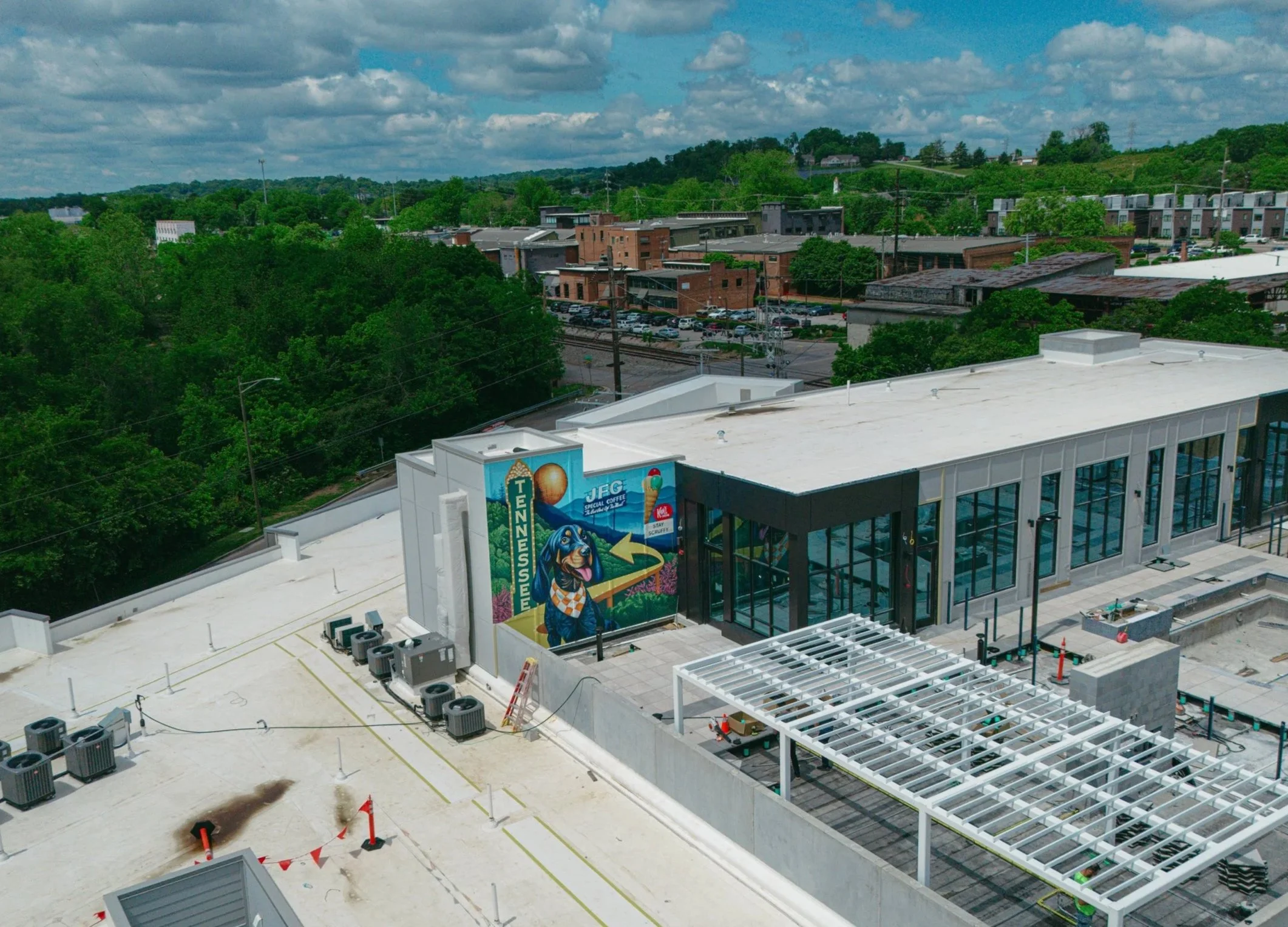 A large building under construction with a mural of Smoky the UT dog, Tennessee marquee on one side. The building has glass windows and a white roof. Aerial photo of The Mark Knoxville