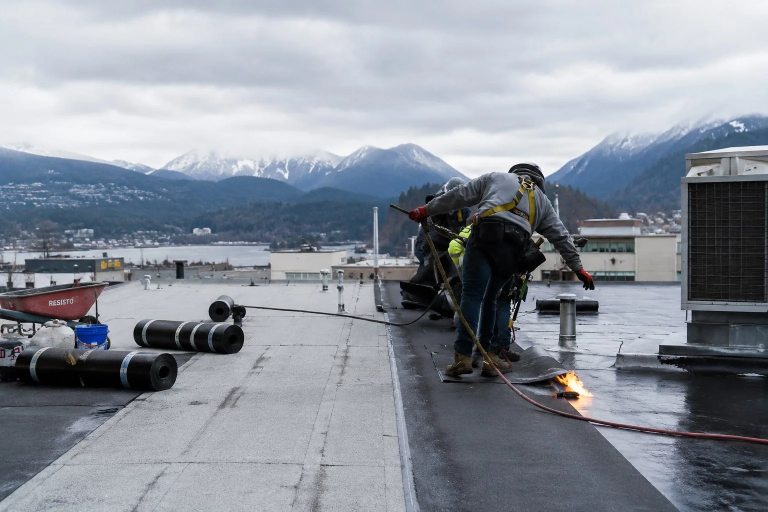 Professional roofing contractors installing a commercial flat roof in West Vancouver, BC with the North Shore mountains in the background. Amex Roofing & Drainage provides commercial roof replacement, repair, waterproofing, and maintenance services.