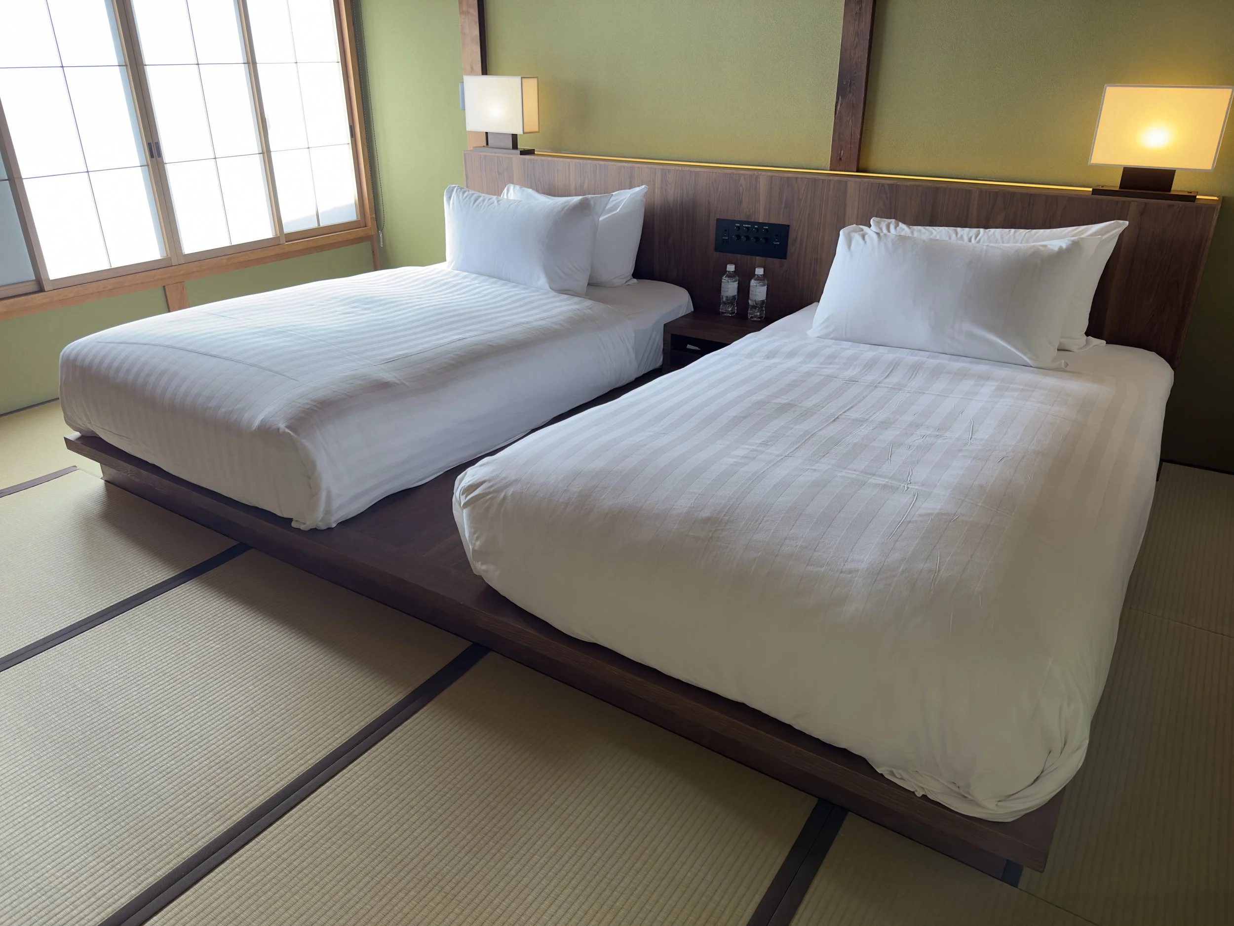 Two neatly made beds with white linens and pillows, positioned on a wooden platform in a room with tatami mat flooring, beside large windows and a green wall.