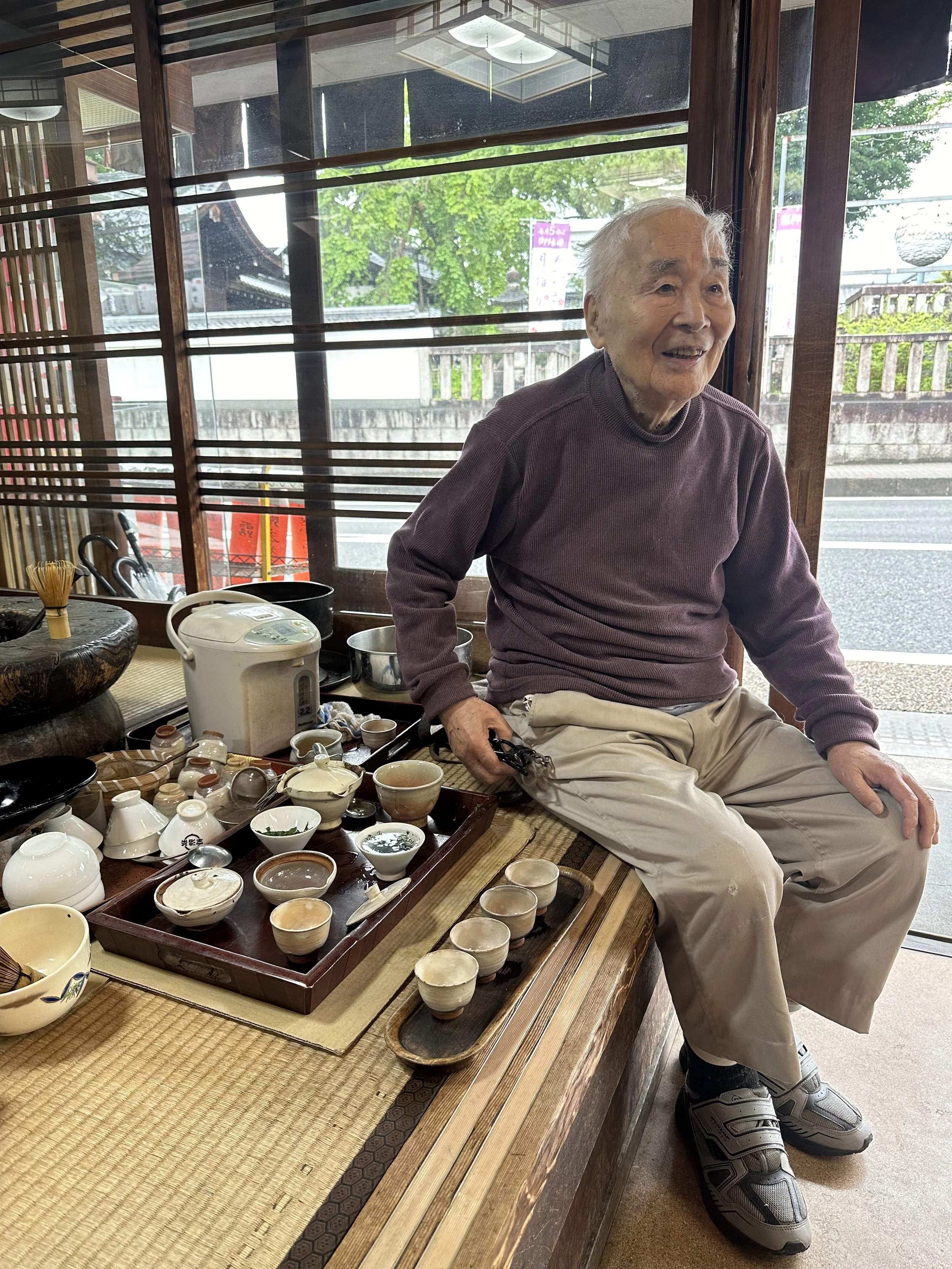 An elderly man sitting on a wooden table inside a traditional Japanese restaurant, surrounded by tea cups, bowls, and a rice cooker, with a window overlooking a street and greenery outside.
