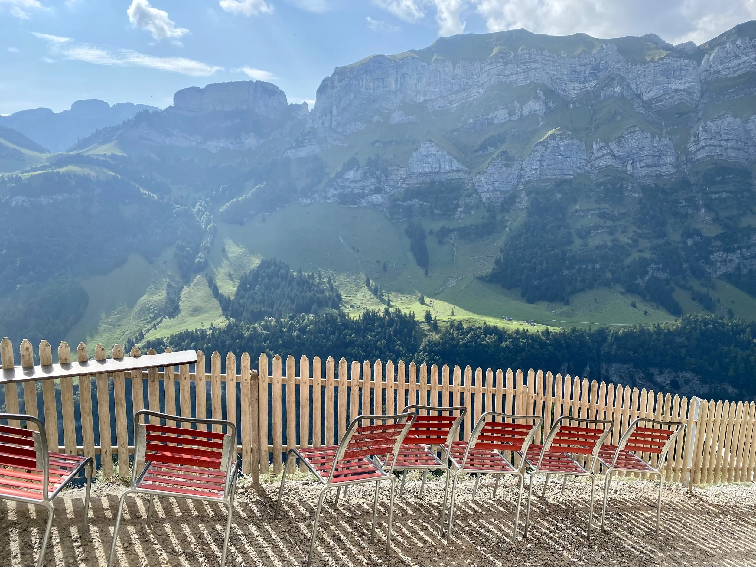 Multiple red and metal chairs on a gravel patio with a wooden fence, overlooking green mountains and blue sky.