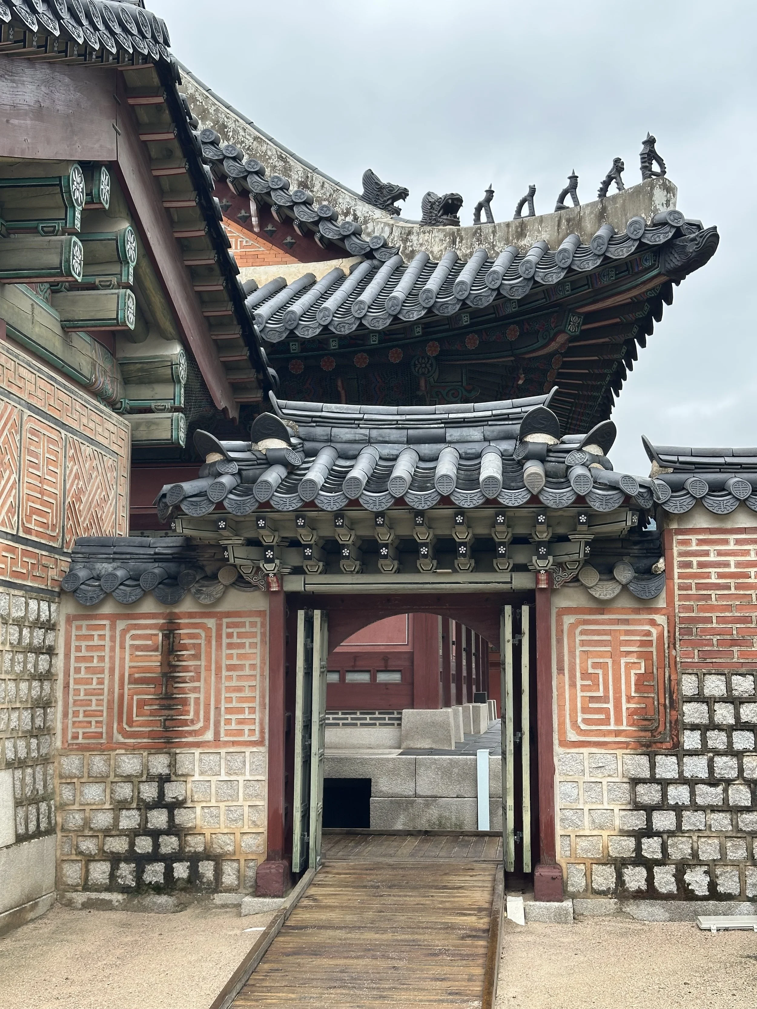 Traditional Asian temple entrance with ornate gray tiled roof, brick and stone walls, and a wooden gate.