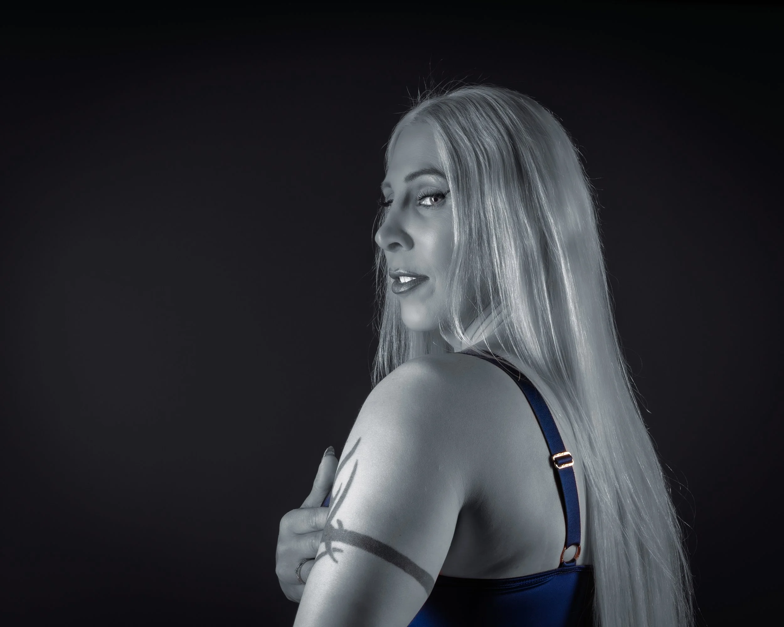 A black and white portrait of a woman with long blonde hair, looking over her shoulder at the camera, wearing a dark tank top with blue straps, with a tattoo on her upper arm, against a black background.